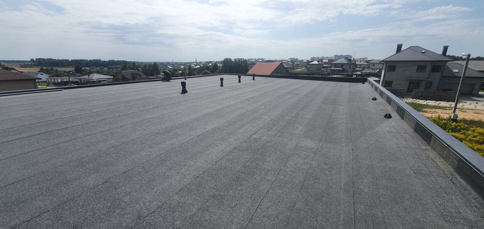 Widok z góry na płaski dach pokryty papą termozgrzewalną, z widocznymi kominkami wentylacyjnymi i obróbkami blacharskimi, w tle zabudowa jednorodzinna i tereny zielone.