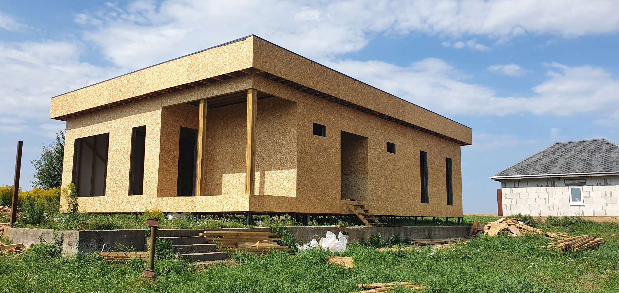 Surowy szkielet domu z płyty OSB na betonowym fundamencie, widoczne schody wejściowe i elementy konstrukcyjne, w tle błękitne niebo z chmurami i drugi budynek w trakcie budowy.