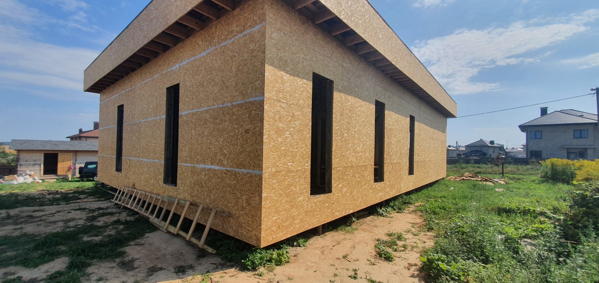 Surowy stan domu szkieletowego obitego płytami OSB z widocznymi otworami okiennymi, na zewnątrz ułożone drewniane schody, w tle inne budynki.