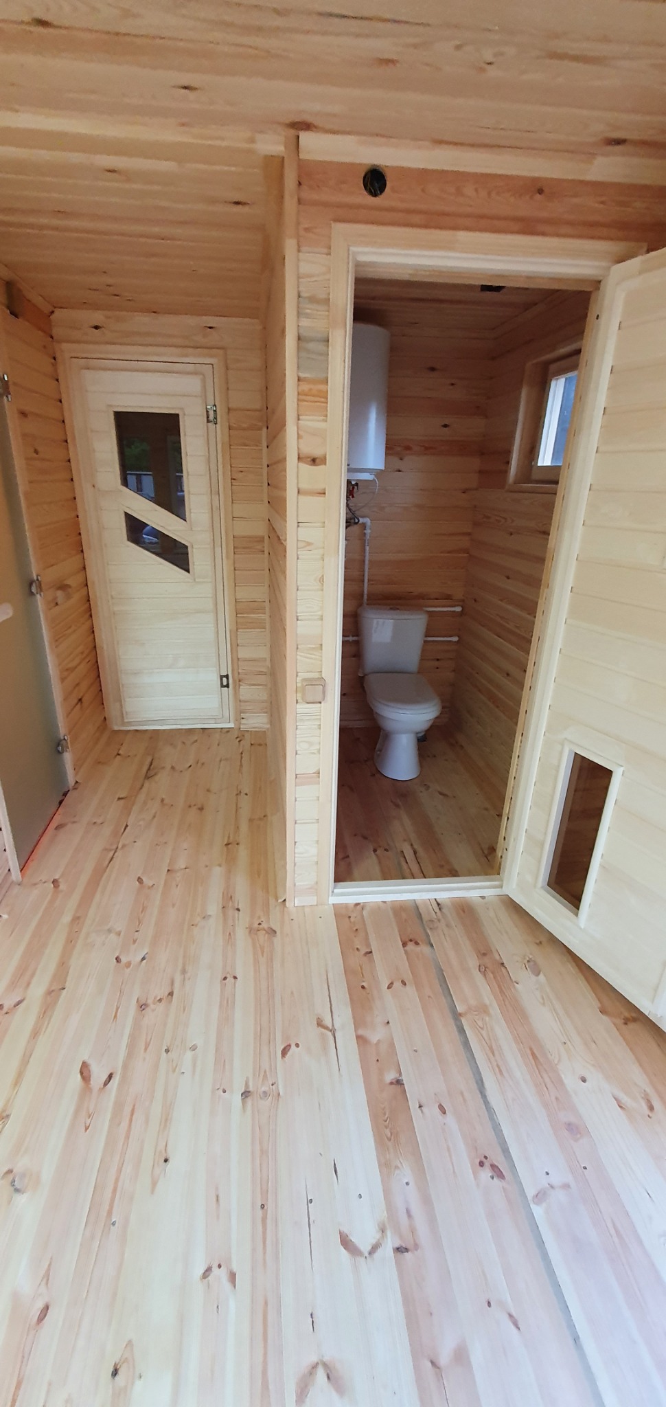 Wnętrze domu szkieletowego z wykończoną łazienką i drewnianą podłogą, widoczna toaleta i bojler w małym pomieszczeniu, oraz drzwi z szybą do innego pokoju.