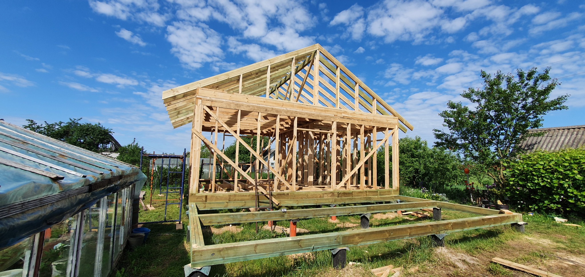 Wznoszenie konstrukcji szkieletowej domu na fundamencie, widoczne drewniane elementy konstrukcyjne i zielone belki fundamentowe, na tle błękitnego nieba z chmurami i zieleni ogrodu.