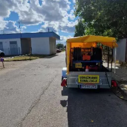Przyczepa z żółtą plandeką i sprzętem do czyszczenia kanalizacji, widoczna tablica z napisem 'Udrażnianie kanalizacji' oraz numer telefonu, na tle budynku i częściowo zachmurzonego nieba.
