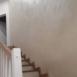 Ściana w neutralnym odcieniu beżu z dekoracyjną strukturą gładzi w pomieszczeniu ze schodami o białych stopniach i drewnianych krawędziach.