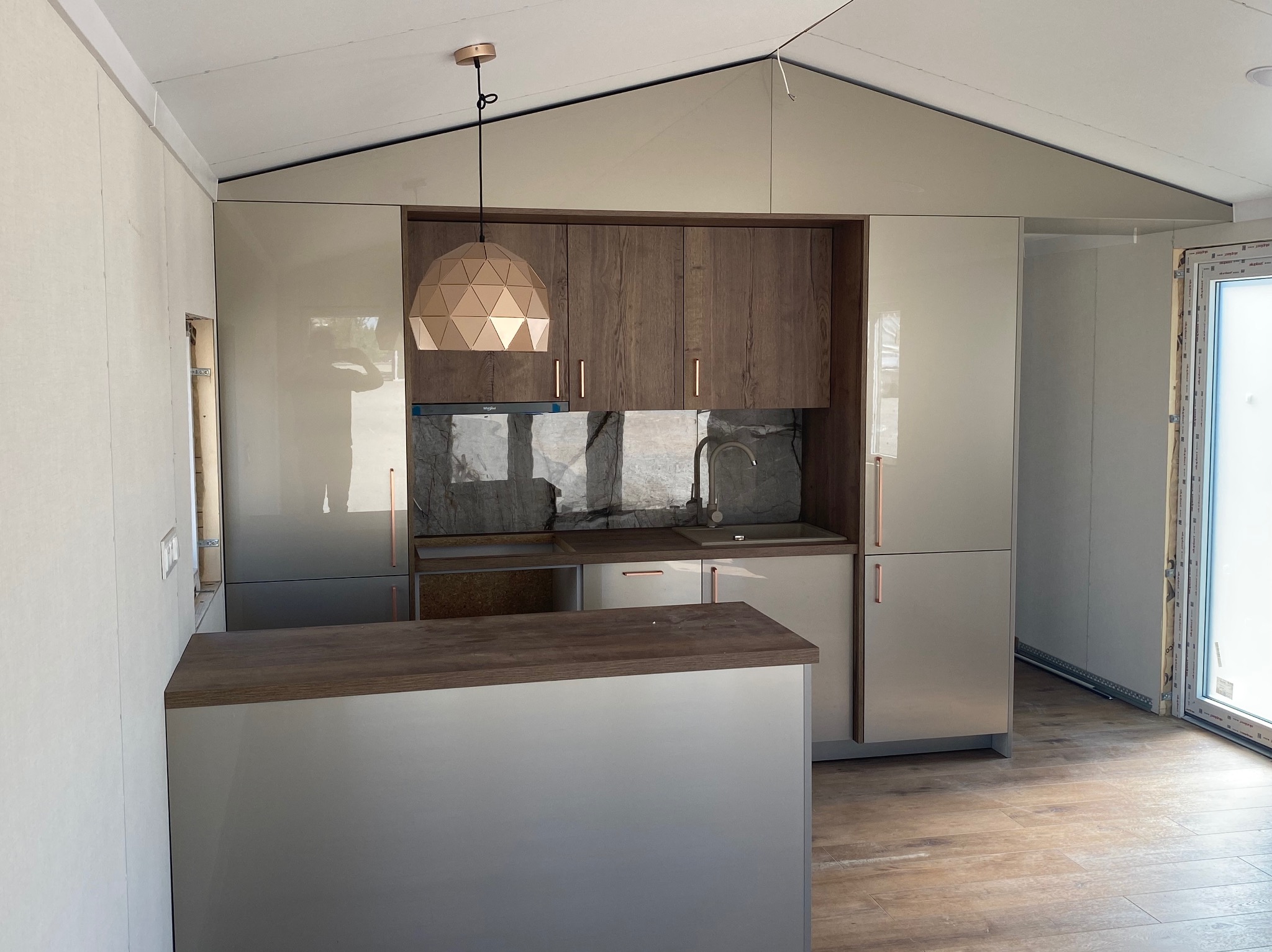 Wnętrze domu z kuchnią w odcieniach beżu i brązu, z drewnianym blatem i geometryczną lampą wiszącą, widok na okno i podłogę imitującą drewno.