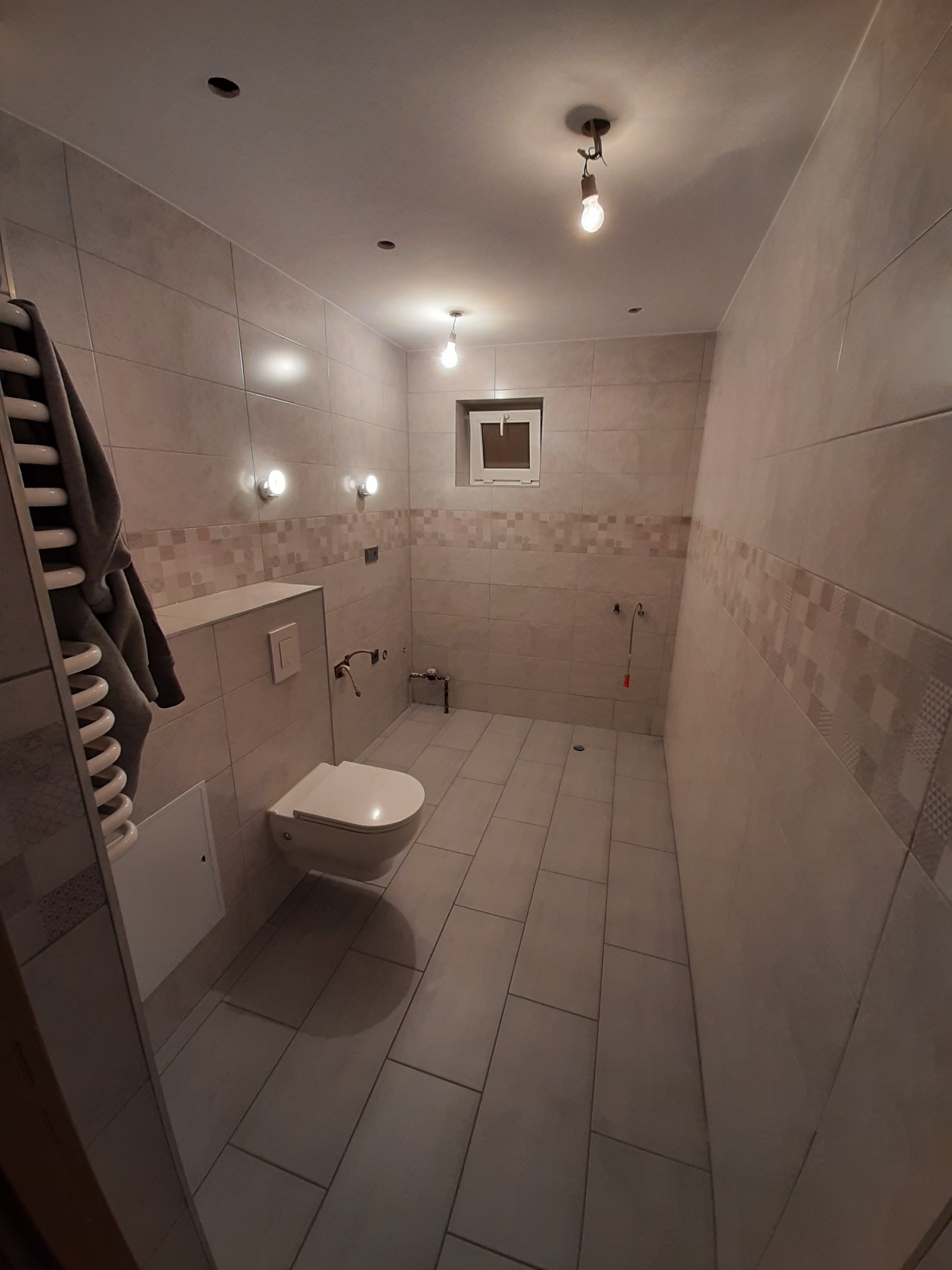 Wąska łazienka w jasnych płytkach z wiszącą toaletą, grzejnikiem drabinkowym z szarym ręcznikiem i surowymi żarówkami na suficie.