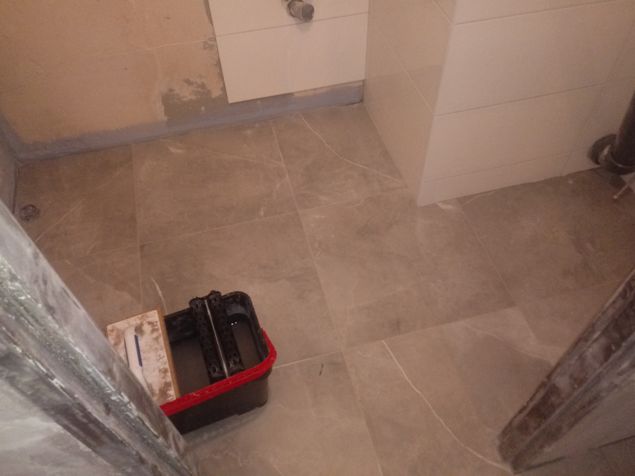 Ułożone płytki podłogowe w łazience, widoczna skrzynka narzędziowa glazurnika i fragment ściany z położonymi płytkami oraz surowa ściana w trakcie prac.