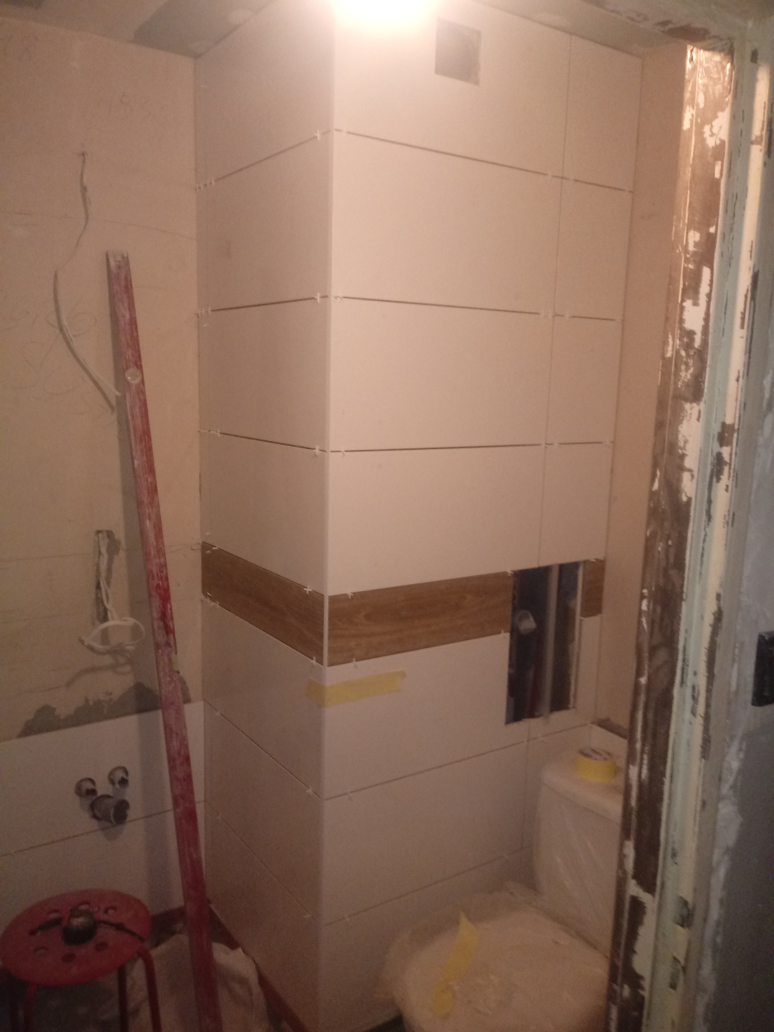 Układanie białych płytek z drewnopodobnym akcentem na narożniku w łazience, widoczna poziomica, taboret i zakryta folią toaleta.