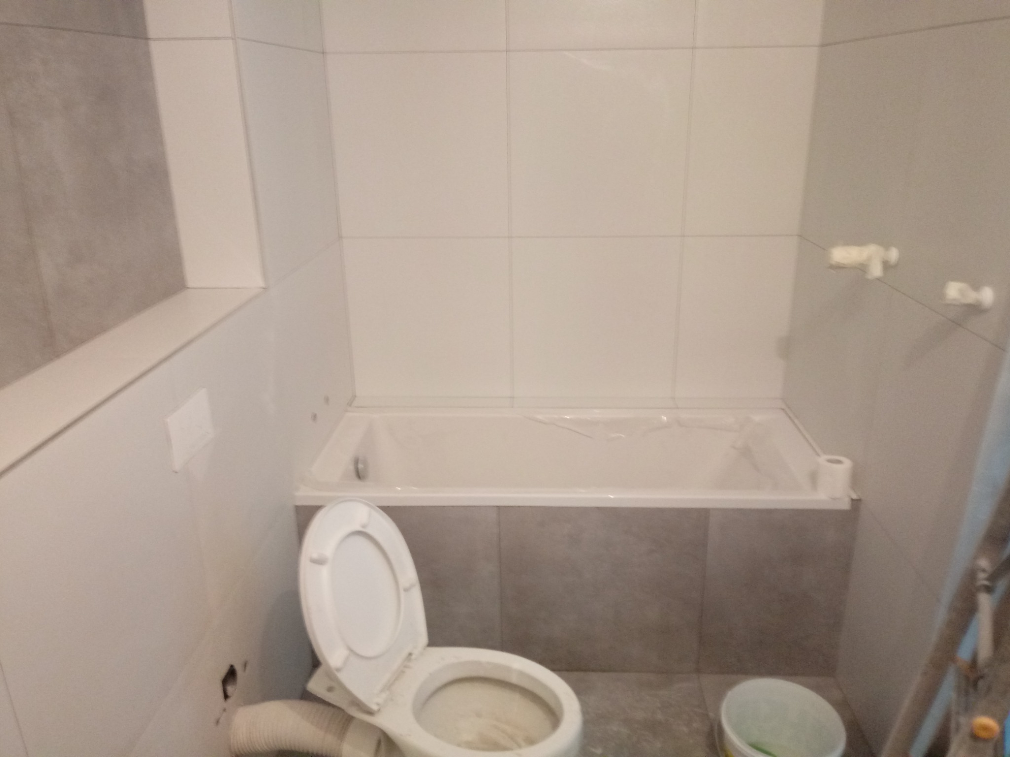 Remontowana łazienka z białą wanną obudowaną szarymi płytkami, częściowo wykończona, widoczne białe płytki na ścianach, biała toaleta z otwartą klapą, drabina malarska i wiadro z farbą wskazują...