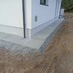 Ułożona kostka brukowa wokół budynku z widoczną skrzynką gazową na elewacji, ślady opon na ziemi.