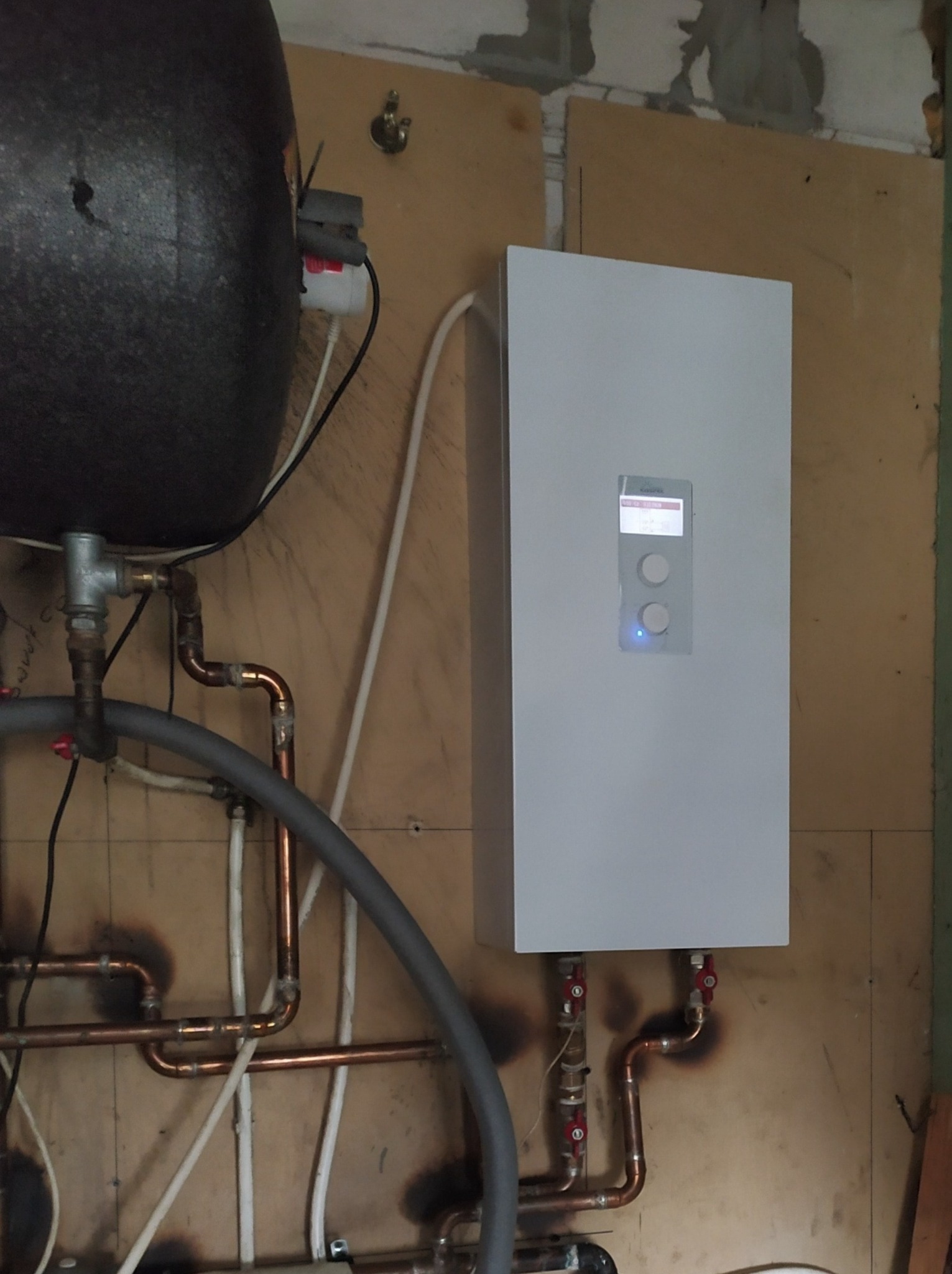 Nowoczesny, biały piec grzewczy wiszący na ścianie, podłączony do instalacji z miedzianych rur. Widoczny zbiornik akumulacyjny obok, z licznymi przewodami. Ściana z widocznymi śladami po remoncie.