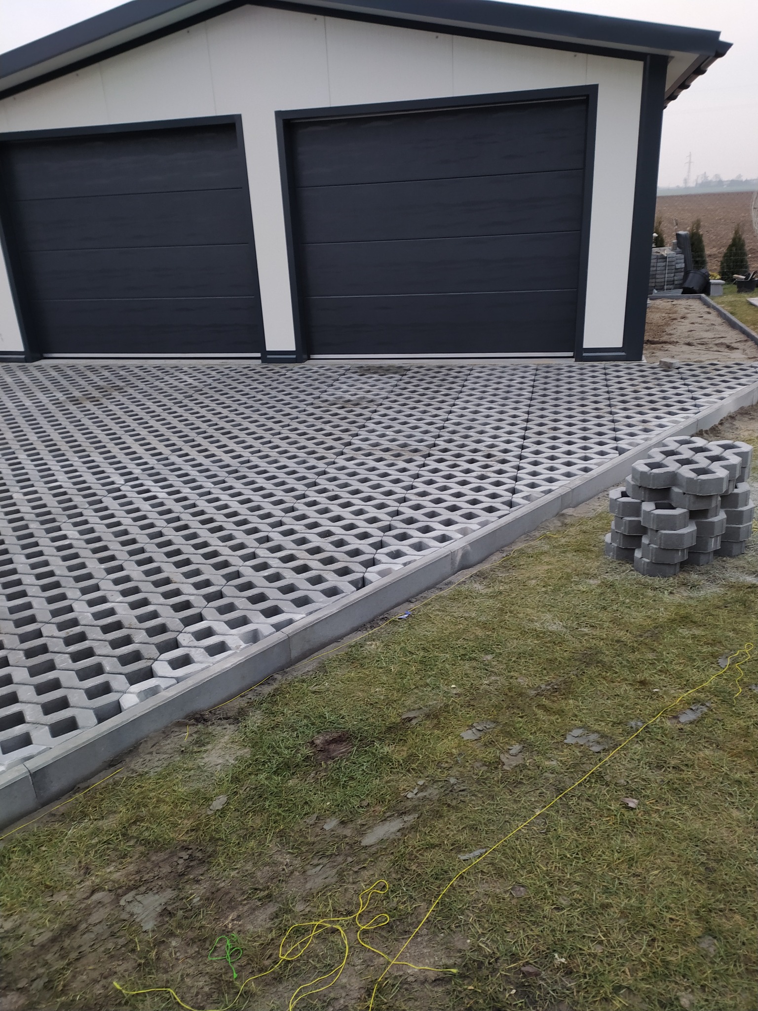 Ułożona ażurowa nawierzchnia z betonowych płyt przed garażem z dwoma bramami, obrzeża wykończone szarym krawężnikiem, obok stos kostki, widoczne linki wyznaczające obszar prac.