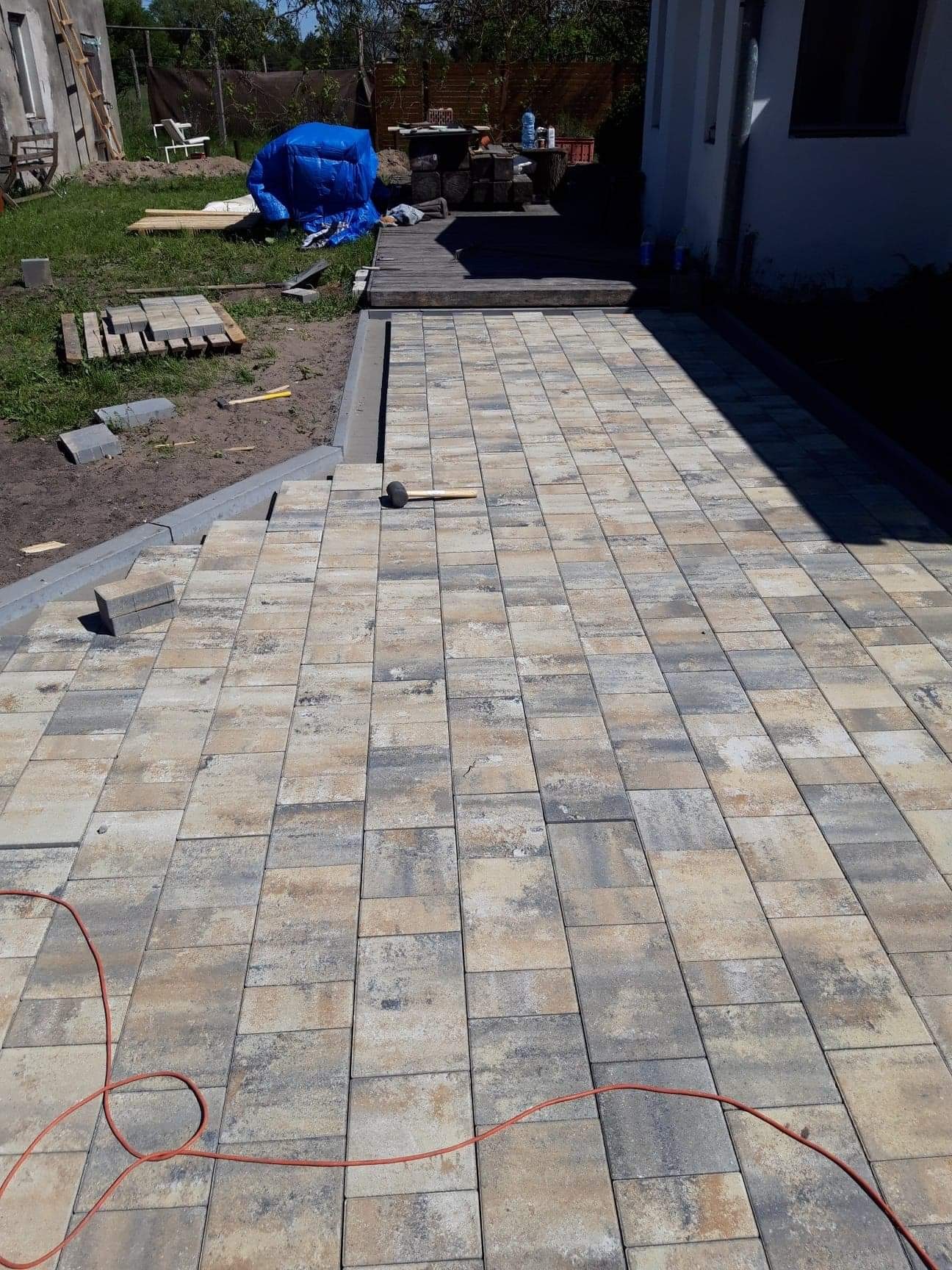 Układanie kostki brukowej z szaro-beżowych elementów; widoczny gumowy młotek do dobijania kostki, paleta z kostką, oraz częściowo ułożona obrzeża.