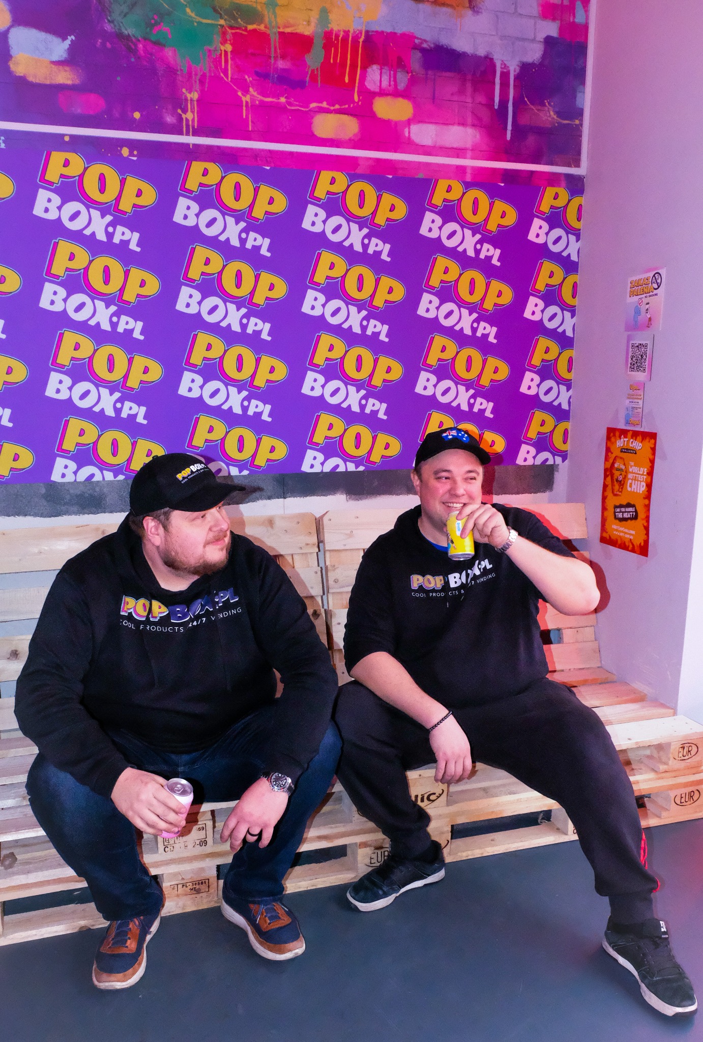 Dwóch mężczyzn w bluzach z logo 'POP BOX' siedzi na paletach i pije napoje w puszkach, w tle ściana z powtarzającym się logo 'POP BOX.PL' i plakat 'HOT CHIP'.