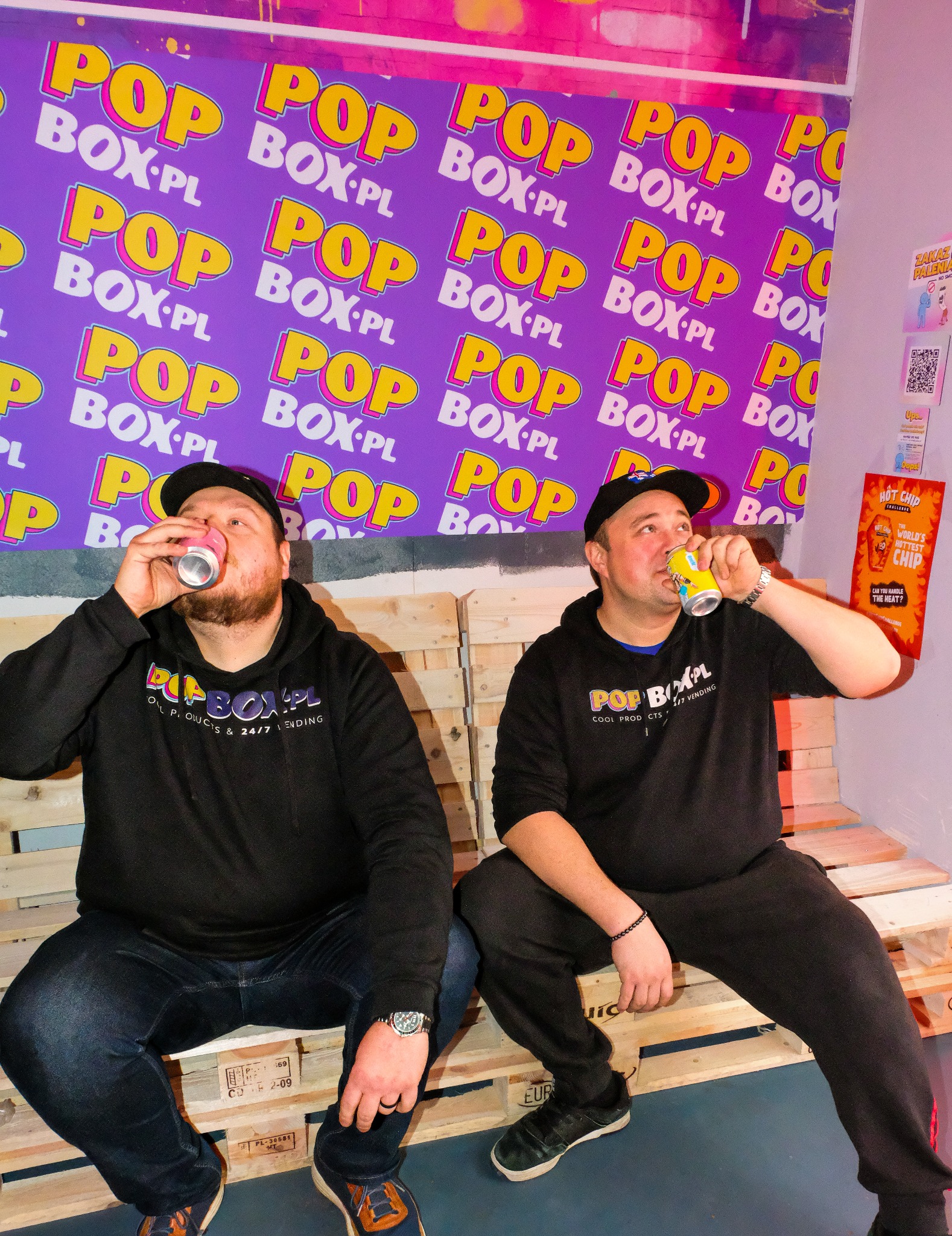 Dwóch mężczyzn w bluzach z logo POP BOX.PL pije napoje z puszek siedząc na paletach, na tle ściany z powtarzającym się logo firmy.