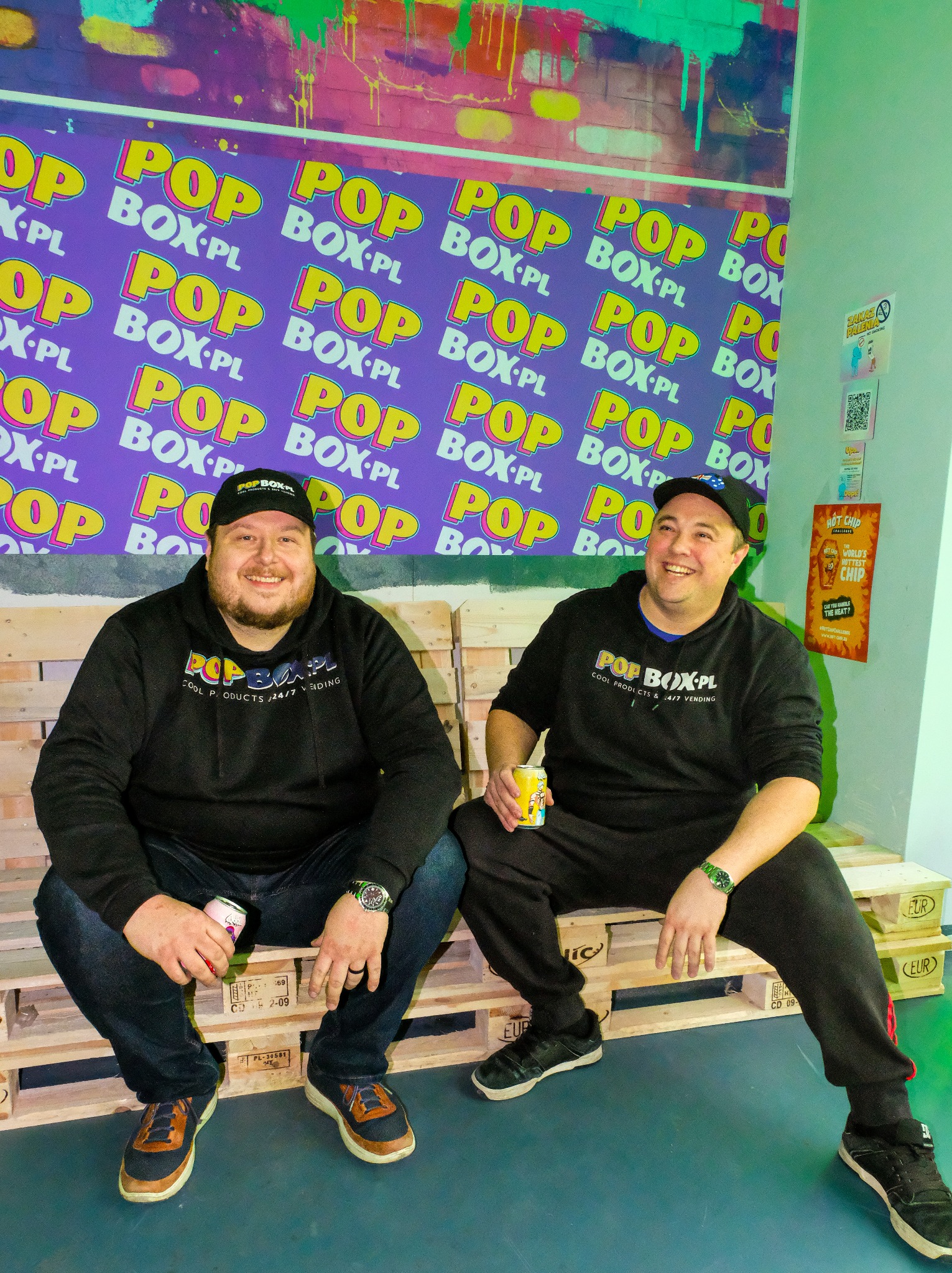 Dwóch mężczyzn w czarnych bluzach z logo POP BOX-PL siedzi na paletach w pomieszczeniu z fioletową ścianą w logo POP BOX-PL, trzymając napoje w puszkach.