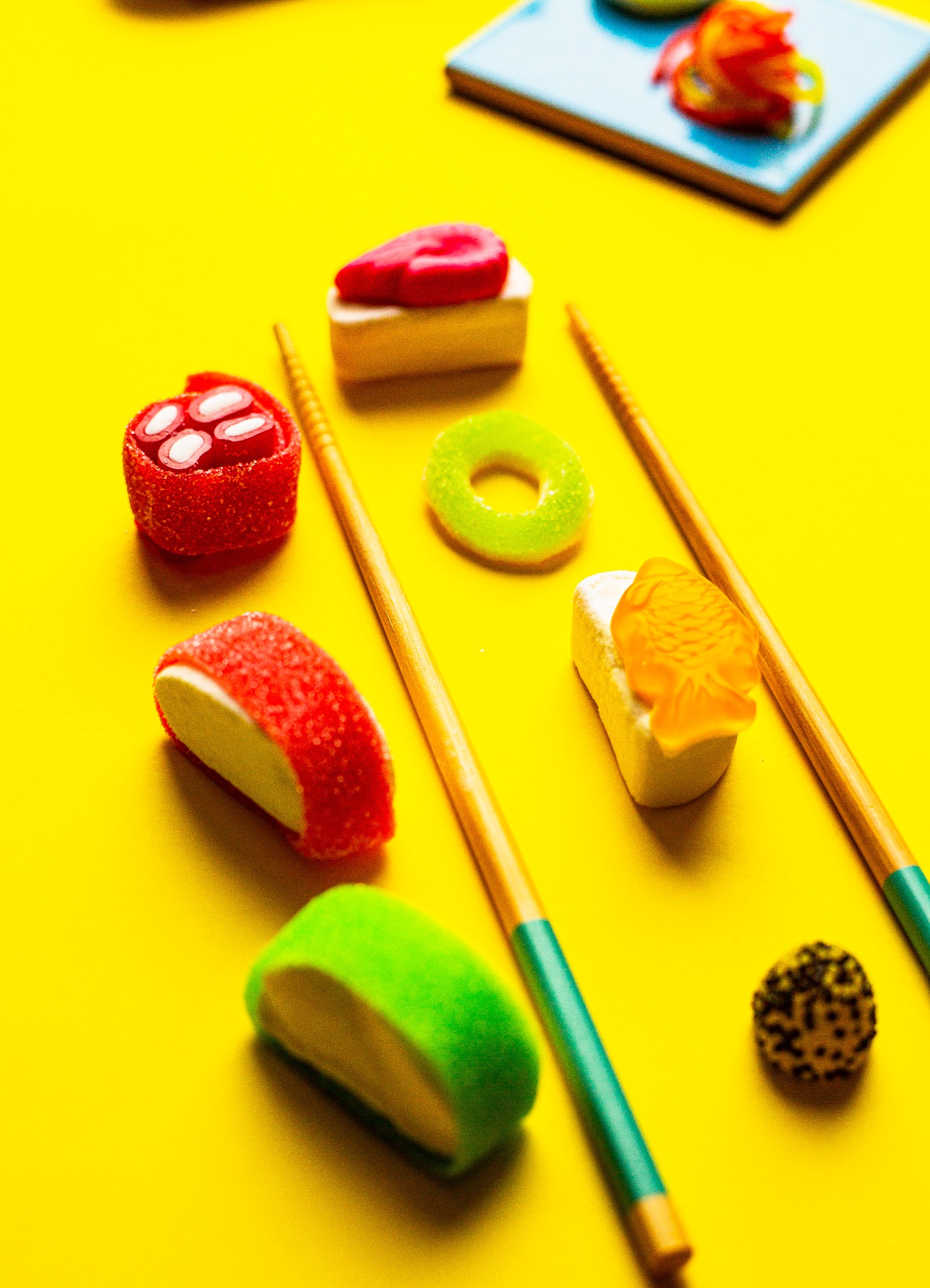 Kolorowe żelki ułożone jak sushi z pałeczkami na jaskrawożółtym tle, imitacja japońskich smaków w słodkiej odsłonie.