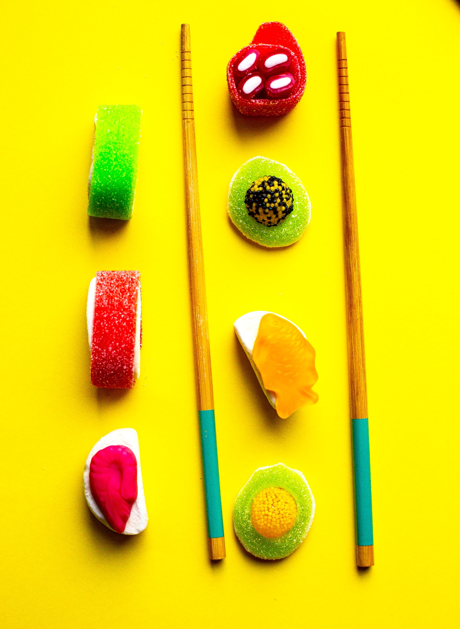 Kompozycja z kolorowych żelków o różnych kształtach i smakach, ułożonych między parami pałeczek do jedzenia na jaskrawożółtym tle, imitująca sushi.