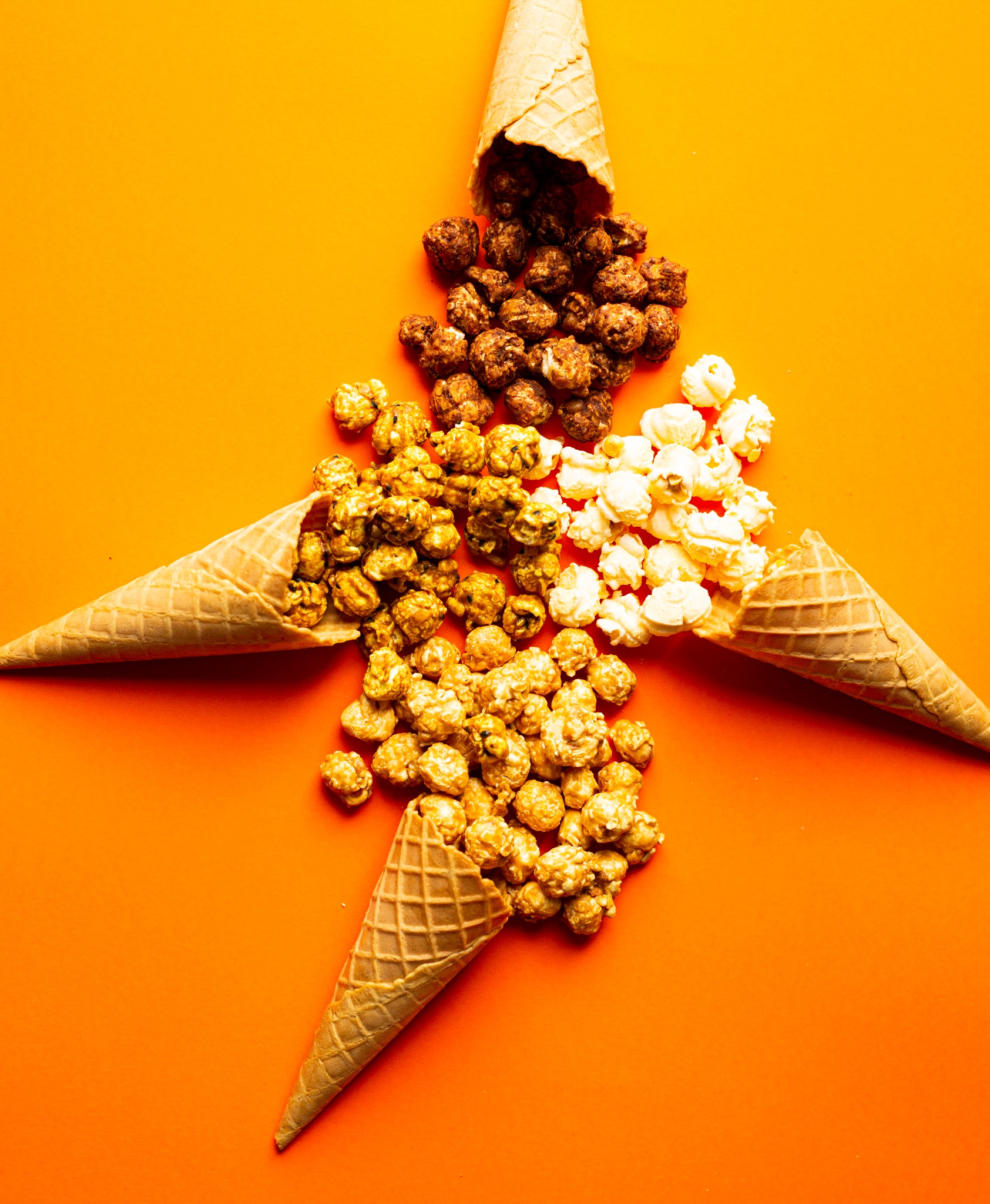 Cztery wafelkowe rożki ułożone w kształt gwiazdy, z których wysypują się różne rodzaje popcornu: karmelowy, czekoladowy i klasyczny, wszystko na jasnopomarańczowym tle.