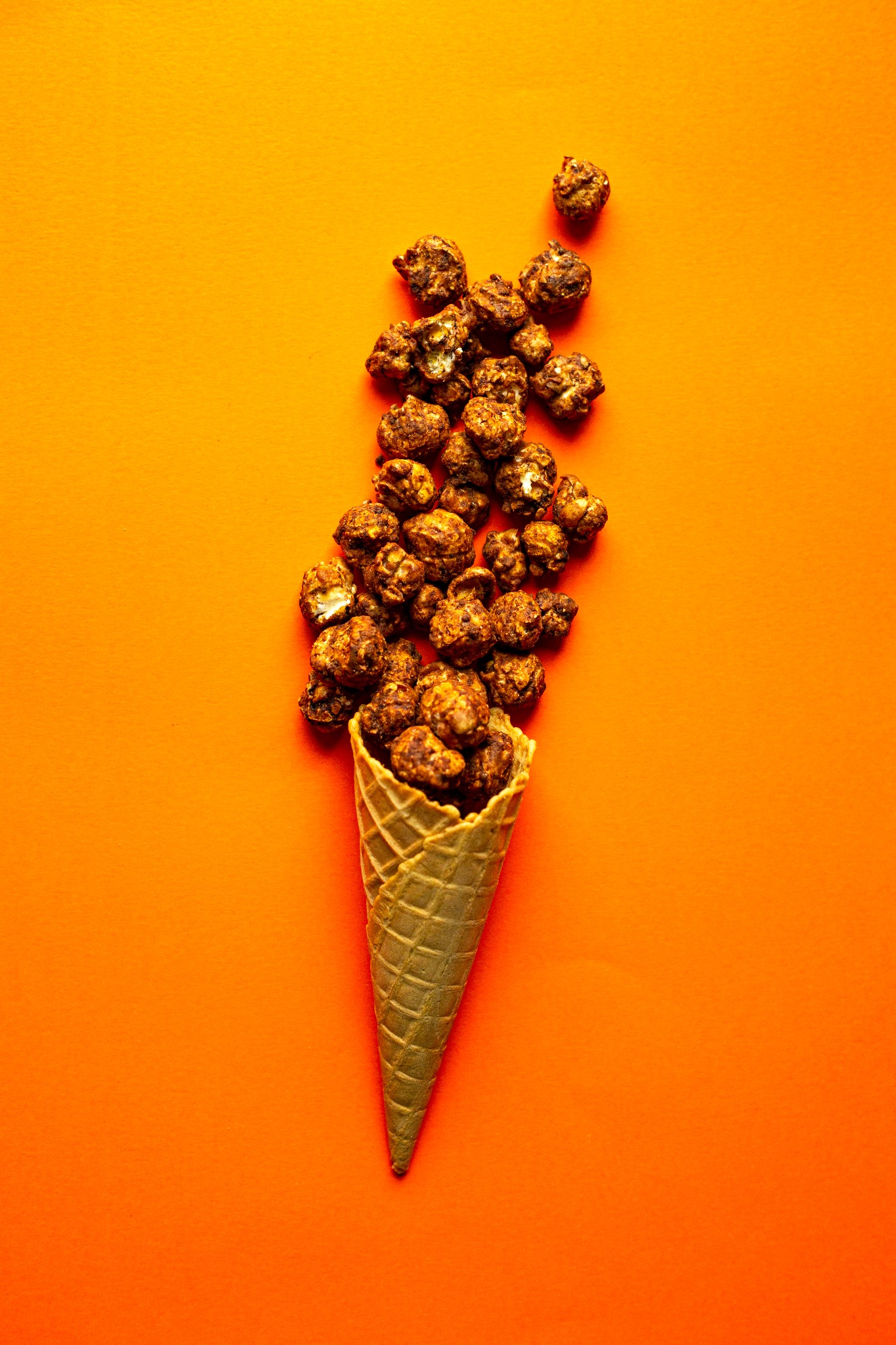 Wafelkowy rożek wypełniony słodkimi, brązowymi kulkami na intensywnie pomarańczowym tle, kilka kulek rozsypanych obok.