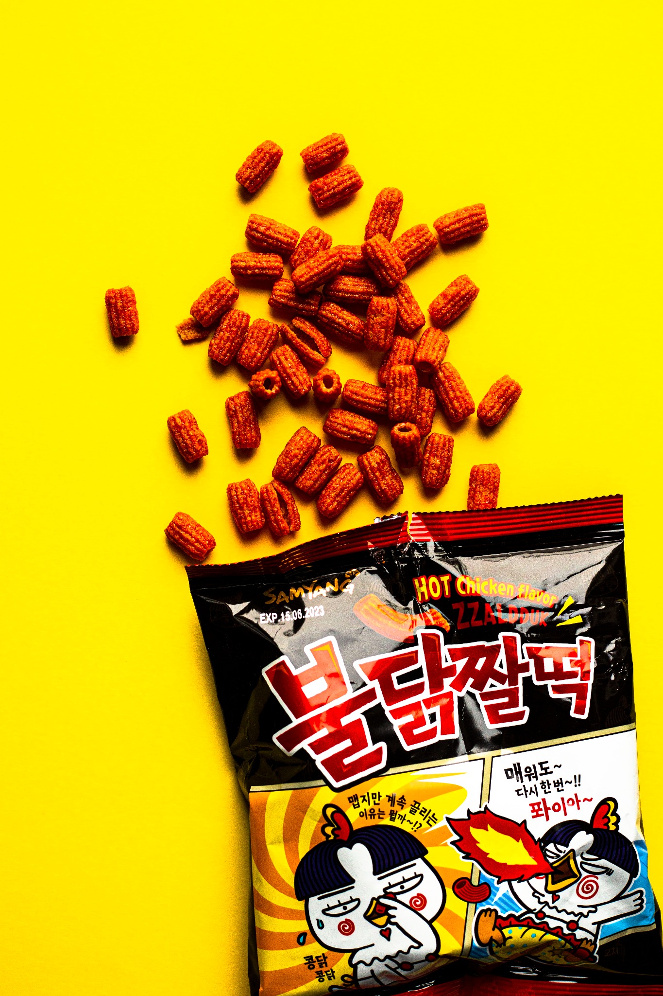 Ostre koreańskie przekąski ryżowe Buldak Zzaldduk rozsypane obok otwartego opakowania na żółtym tle, widoczny termin ważności 15.06.2023 i grafika z kurczakiem.