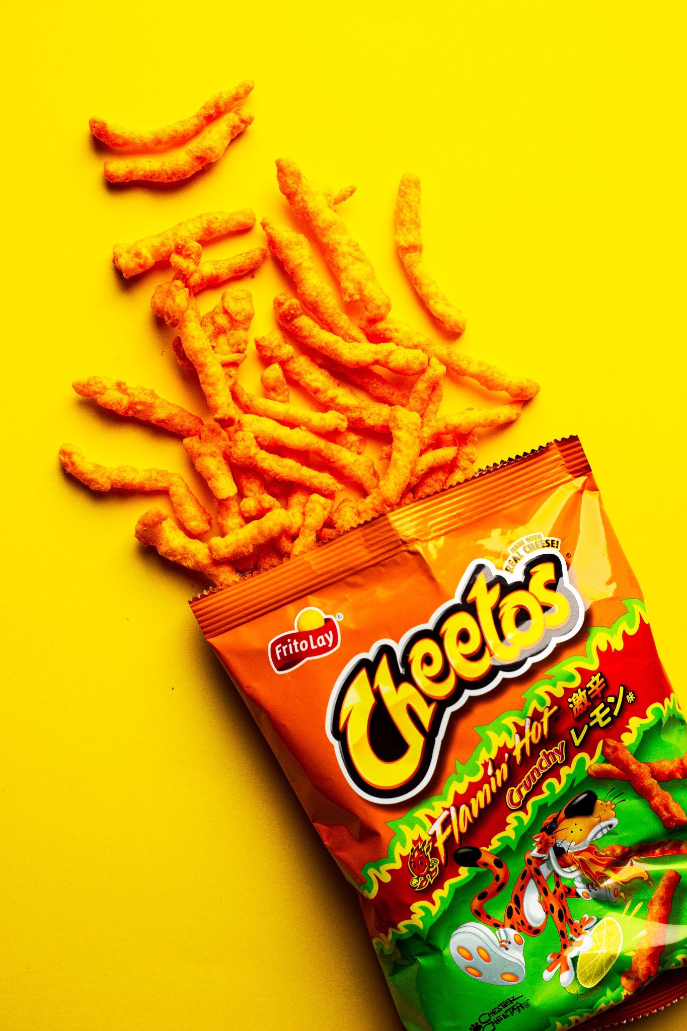 Rozsypane pomarańczowe chrupki Cheetos Flamin' Hot obok otwartej paczki na jaskrawożółtym tle, z widocznym logo Frito Lay i postacią Cheetosa z cytryną.