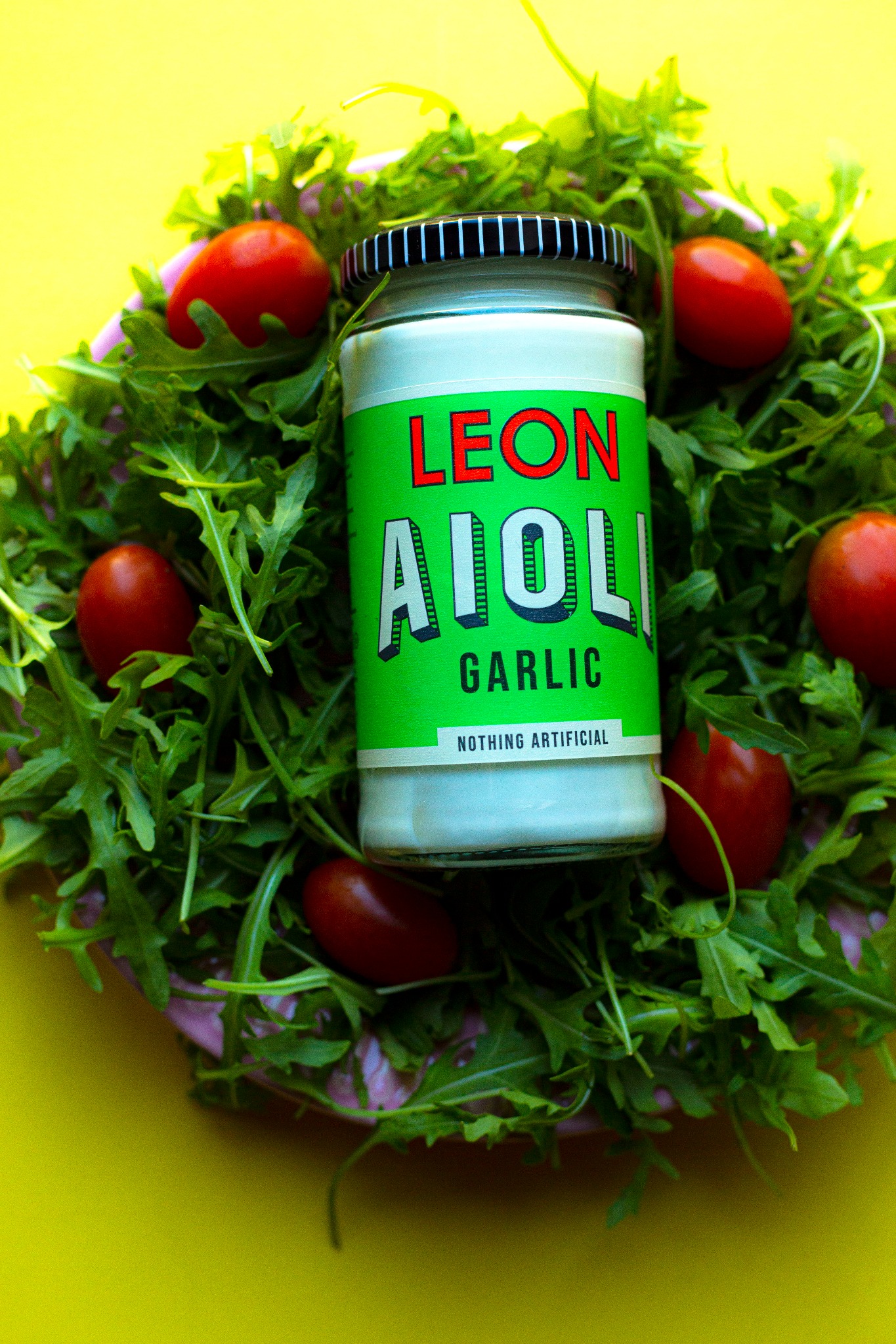 Słoik aioli czosnkowego marki Leon otoczony rukolą i pomidorkami cherry na talerzu z żółtym tłem.
