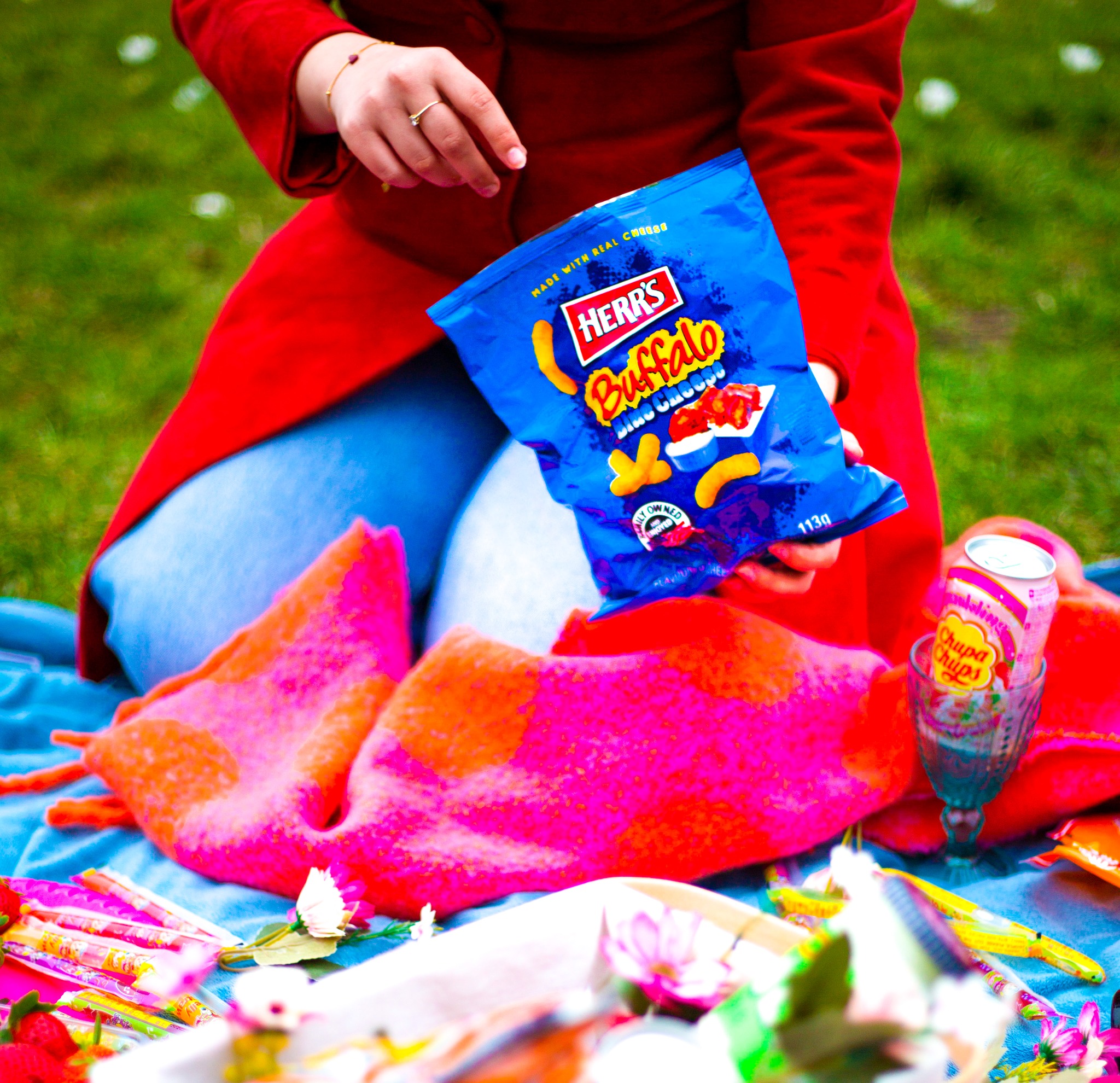 Kobieta w czerwonym płaszczu trzyma paczkę chipsów Buffalo BBQ na pikniku z kocem, napojem Chupa Chups i słodyczami.