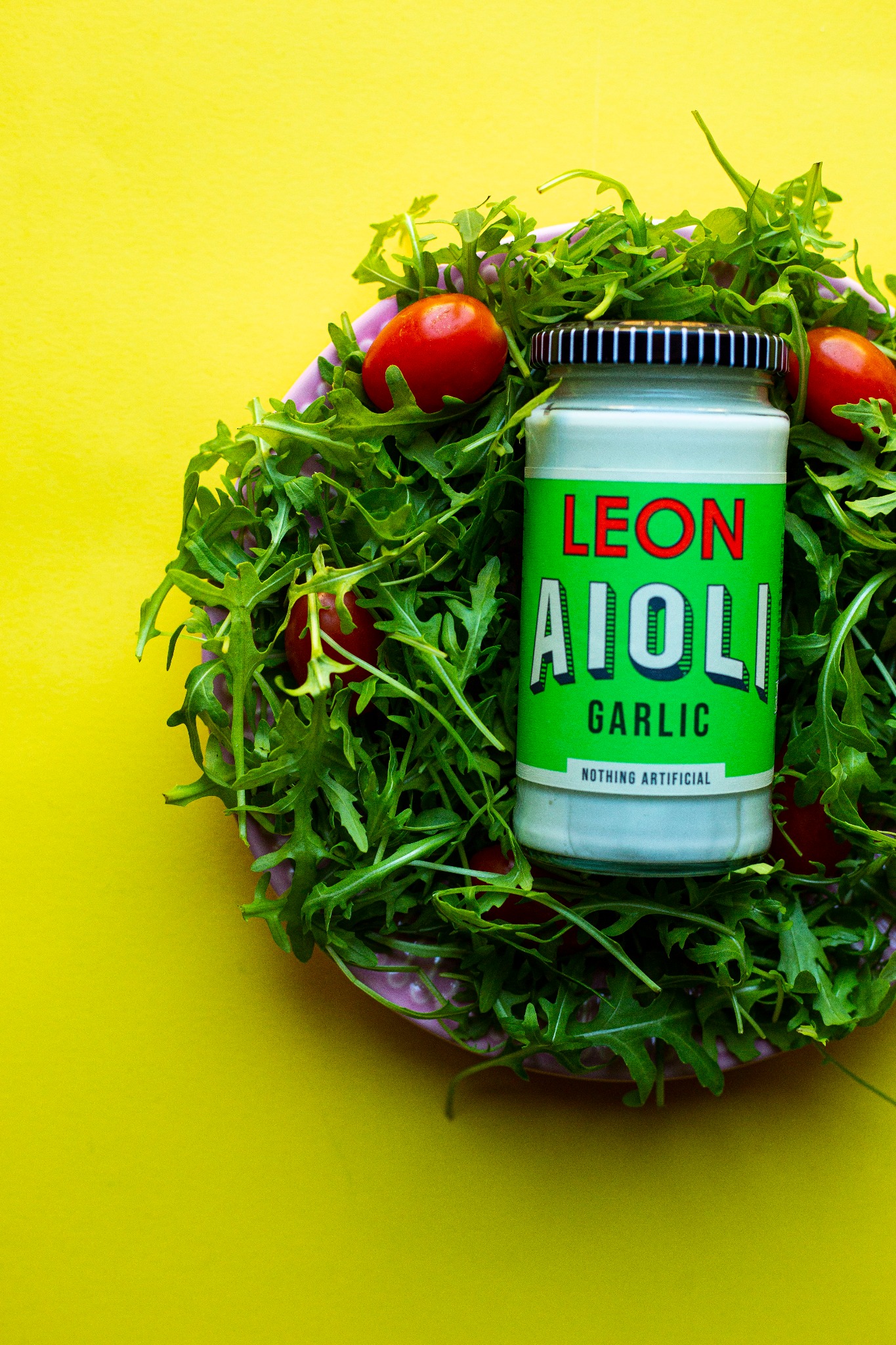 Słoik Aioli marki Leon otoczony rukolą i pomidorkami koktajlowymi na talerzu, na jaskrawożółtym tle.