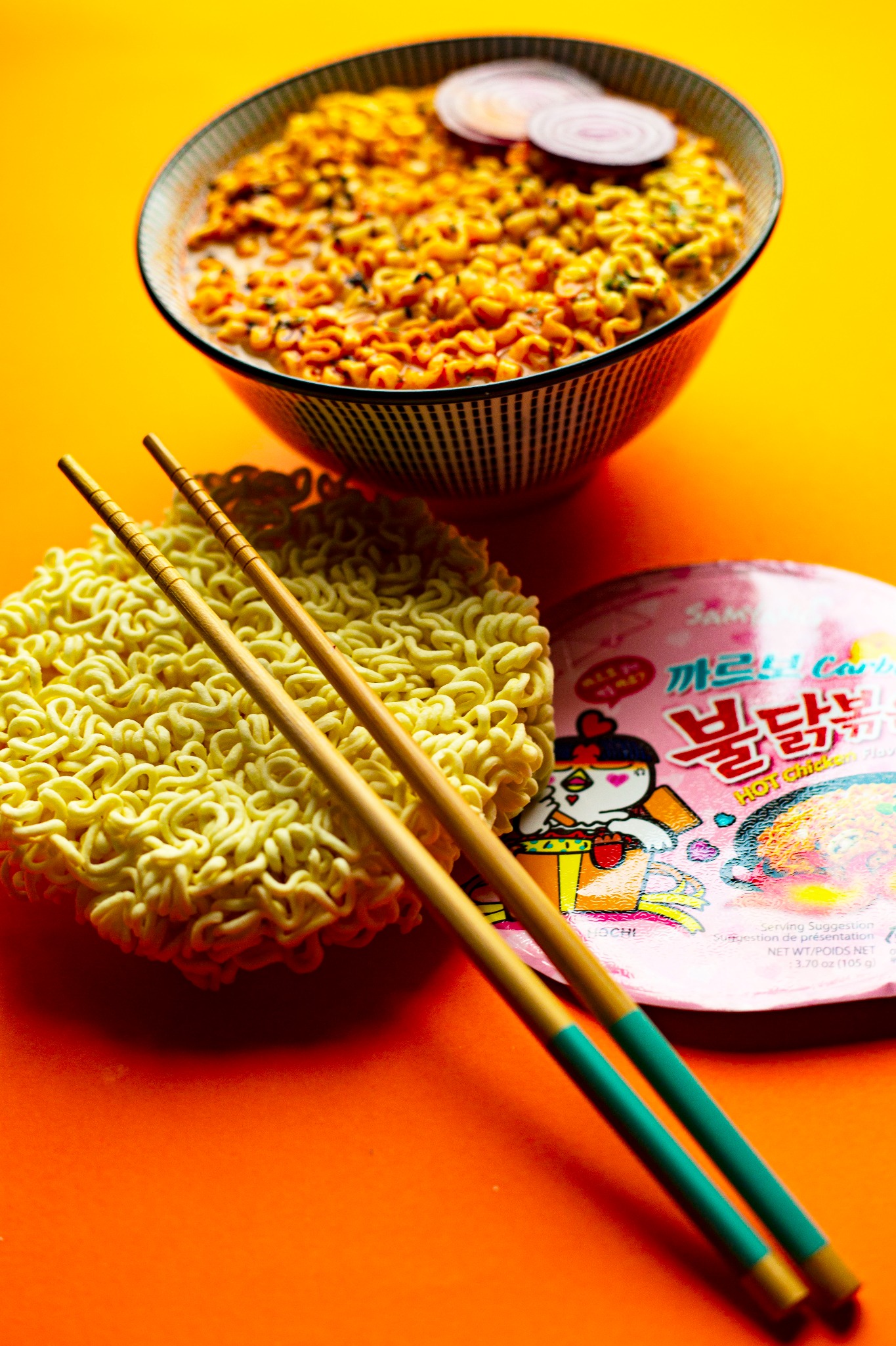 Miseczka pikantnego, koreańskiego makaronu ramen z plasterkami czerwonej cebuli, obok suchego makaronu, opakowanie z wizerunkiem kurczaka i pałeczki z zielonymi uchwytami na pomarańczowym tle.