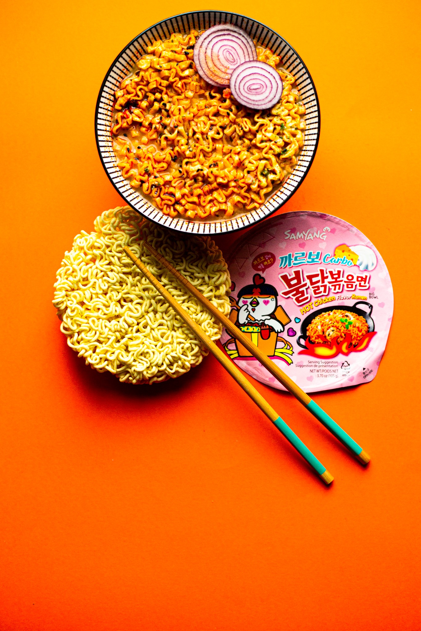 Gotowy makaron instant w misce z dwoma krążkami czerwonej cebuli, obok surowy makaron i opakowanie, pałeczki do jedzenia na pomarańczowym tle.