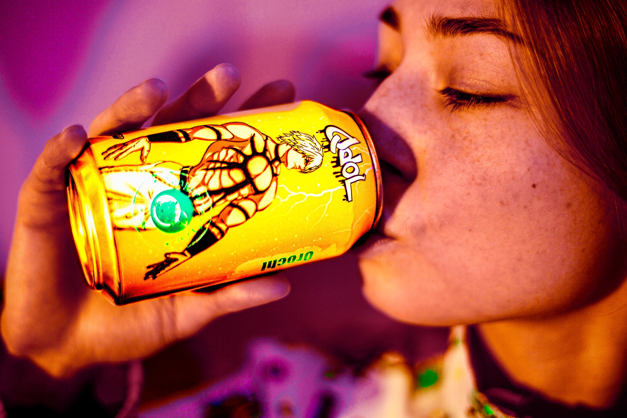 Kobieta pije napój energetyczny z żółtej puszki z wizerunkiem postaci w czarno-pomarańczowym kostiumie i napisem 'Orochi' w odblaskowym oświetleniu.