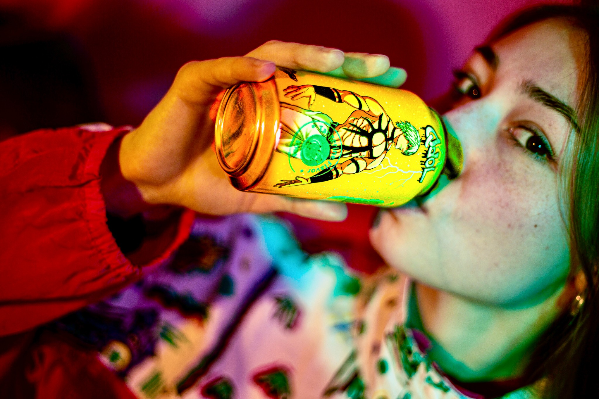 Młoda osoba pije napój z żółtej puszki z rysunkiem postaci w stylu komiksowym, widoczne piegi na twarzy, w tle czerwone i zielone światło.