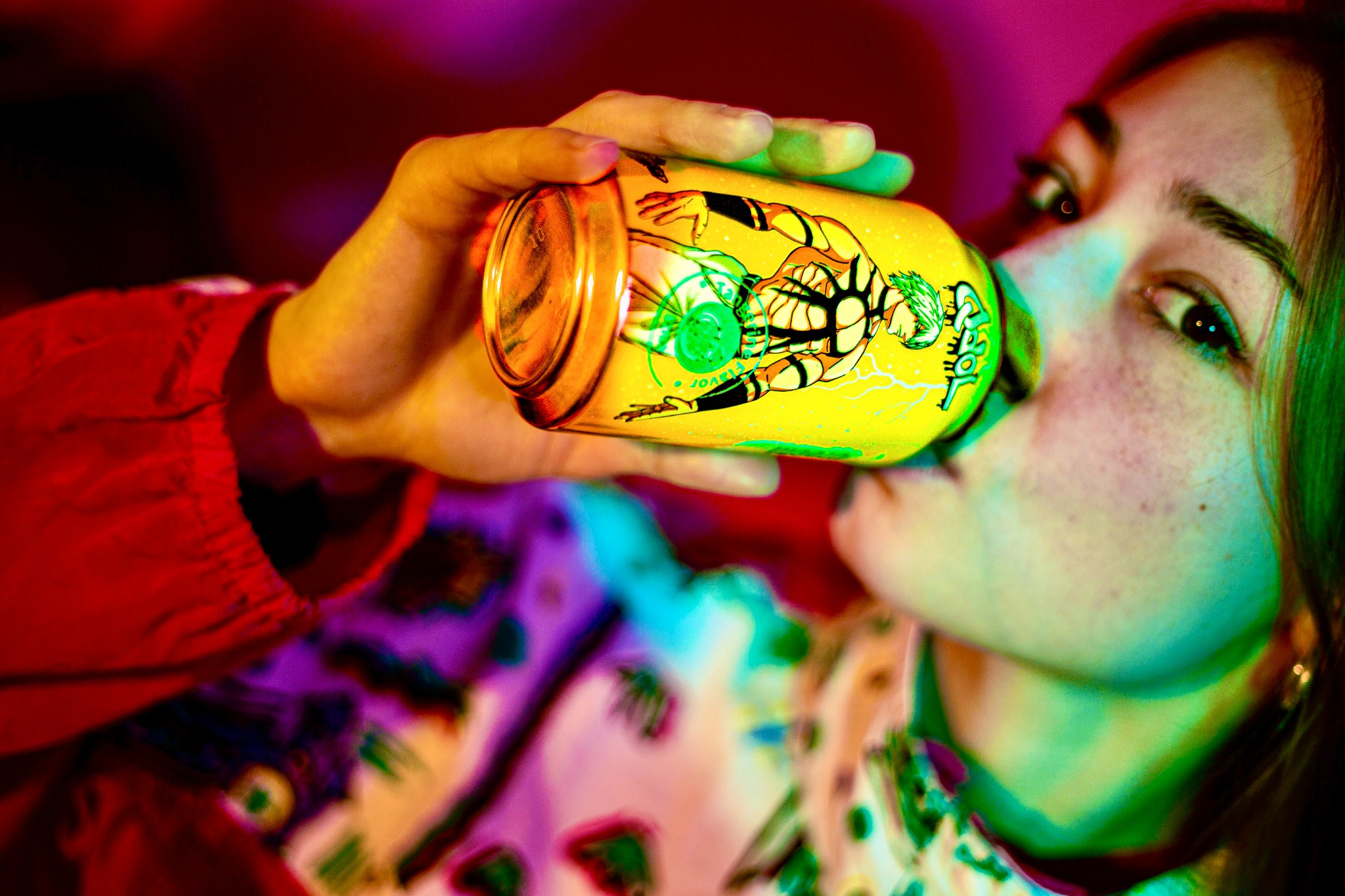 Młoda kobieta pije napój z żółtej puszki z grafiką postaci w stylu anime, oświetlona neonowym światłem.