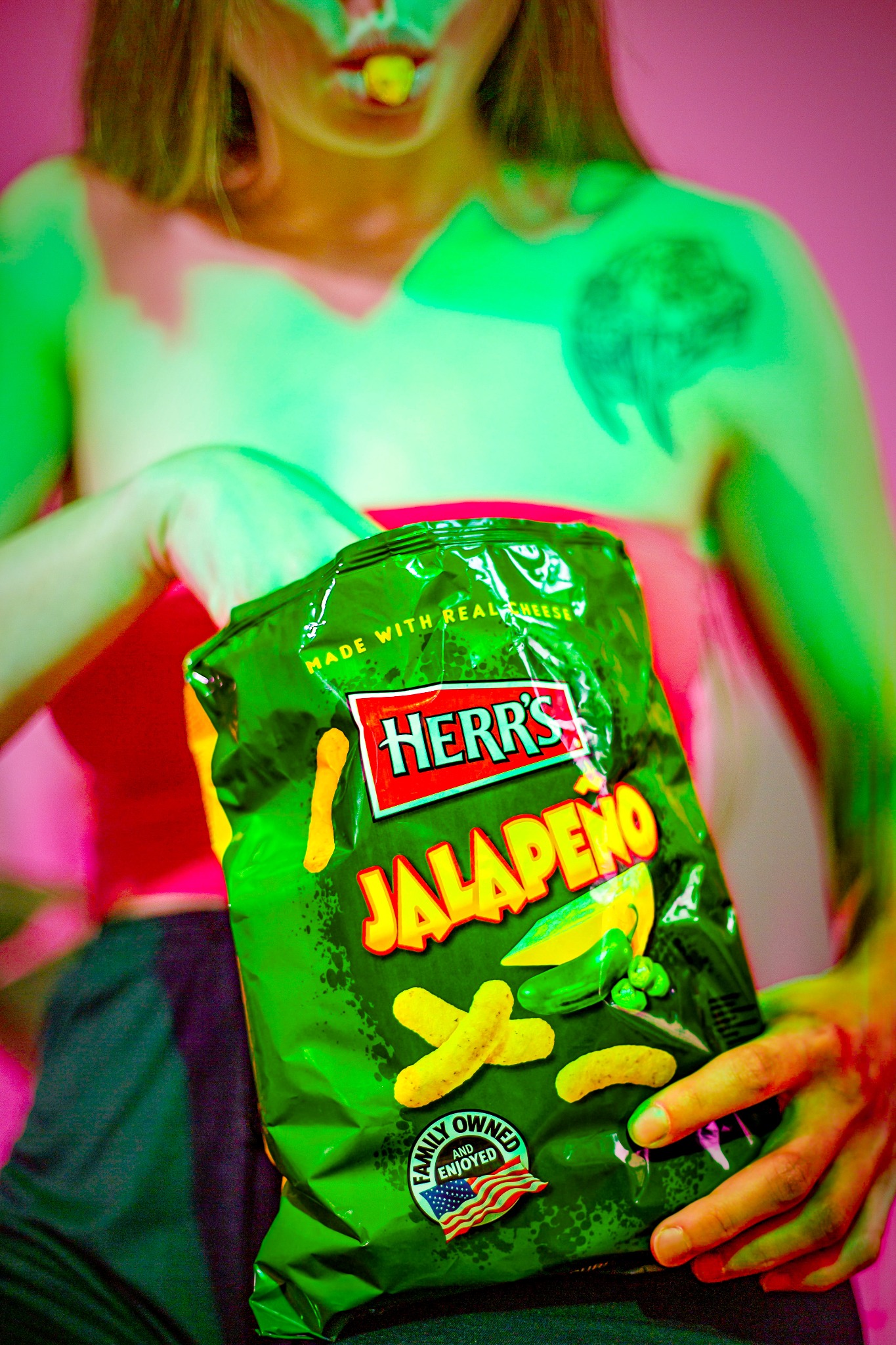 Osoba trzyma zieloną paczkę chrupek o smaku jalapeno, z jednym chrupkiem w ustach i tatuażem na ramieniu, oświetlona intensywnym różowo-zielonym światłem.