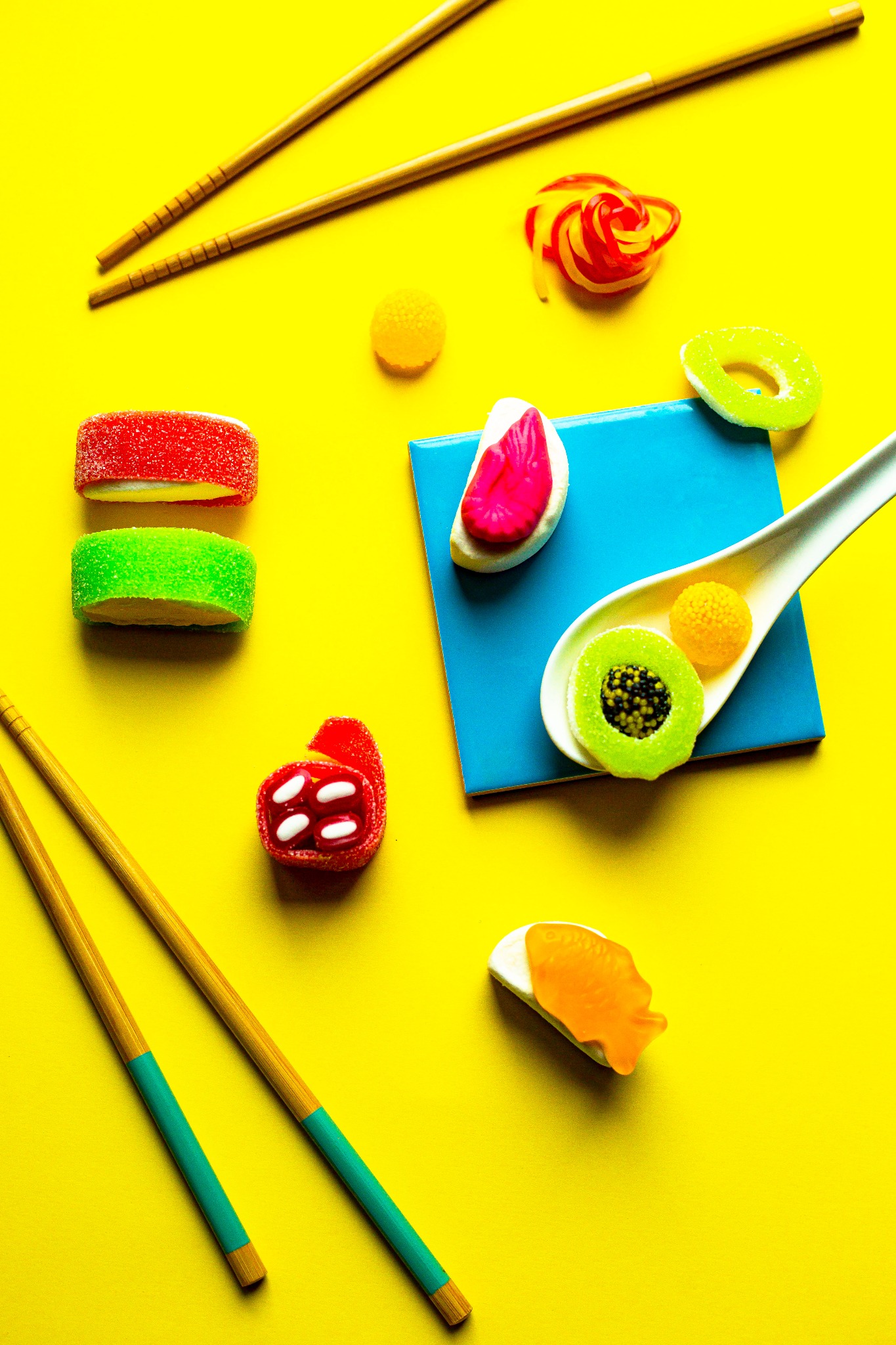Kolorowe żelki ułożone na żółtym tle z pałeczkami do sushi i niebieską płytką, przypominające słodkie sushi.