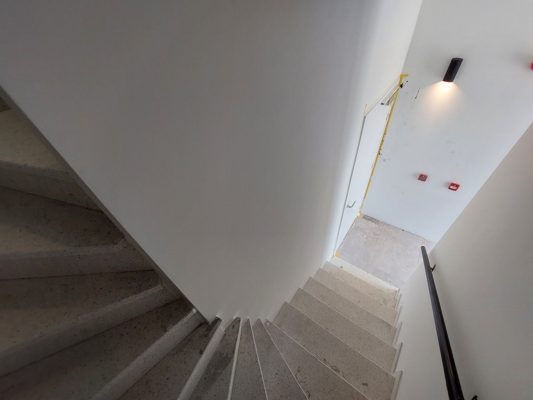 Wnętrze klatki schodowej po malowaniu ścian na biało, widok z góry na schody z lastryko, czarna lampa sufitowa i zabezpieczone taśmą malarską ościeżnice drzwi.