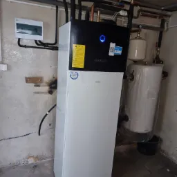 Nowa, biało-czarna pompa ciepła Daikin, jeszcze częściowo w folii ochronnej, zainstalowana w piwnicy z widocznymi rurami, zbiornikiem wyrównawczym i skrzynką elektryczną na ścianie.