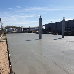 Agbudcompany - Świeżo wylana, gładka betonowa płyta fundamentowa z wystającymi prętami zbrojeniowymi w filarach, widok na plac budowy i błękitne niebo.