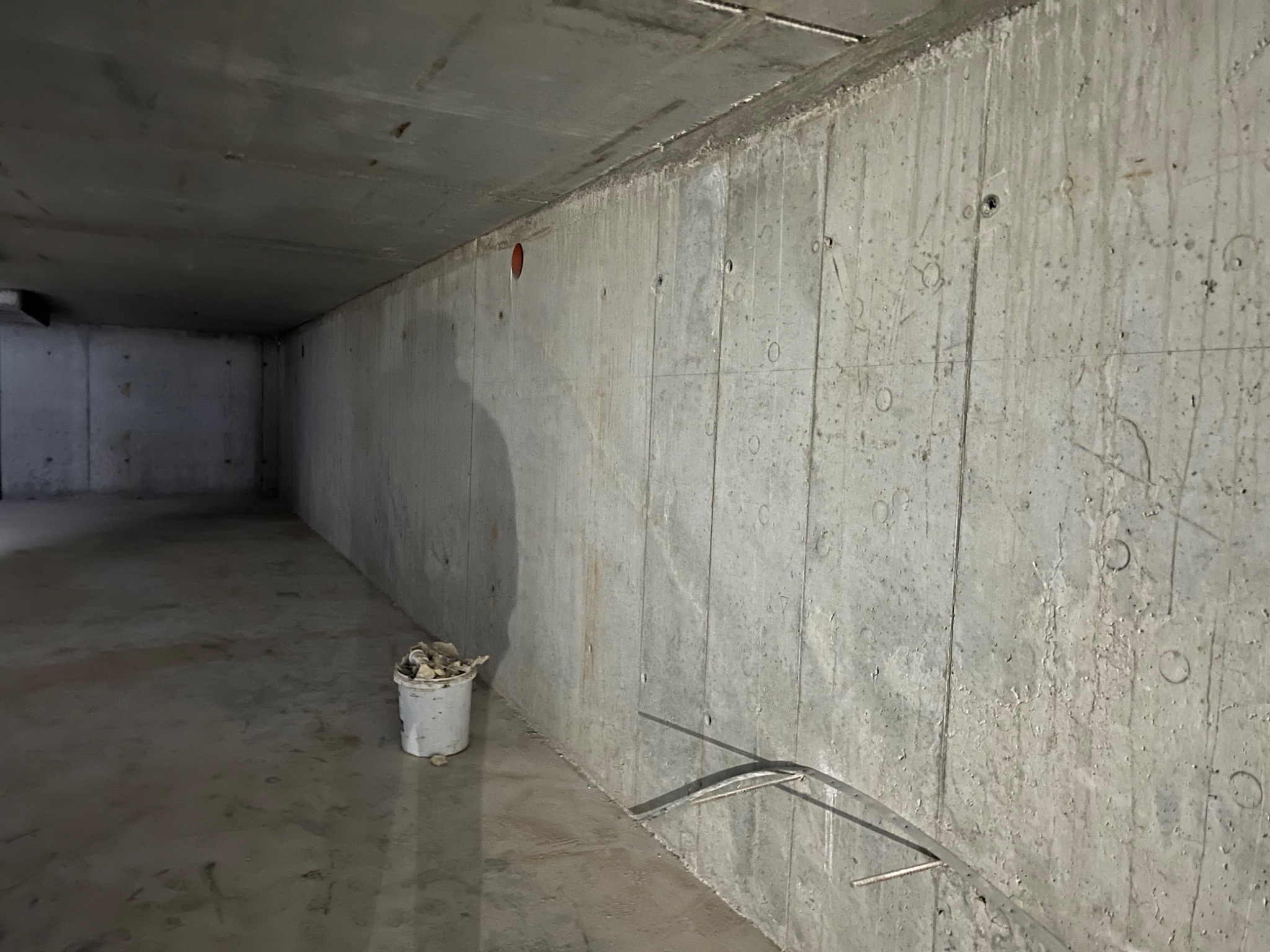 Surowa, betonowa ściana w trakcie budowy, widoczne ślady po szalunkach, mały, biały kubeł z resztkami betonu przy ścianie, wystające pręty zbrojeniowe z podłoża.