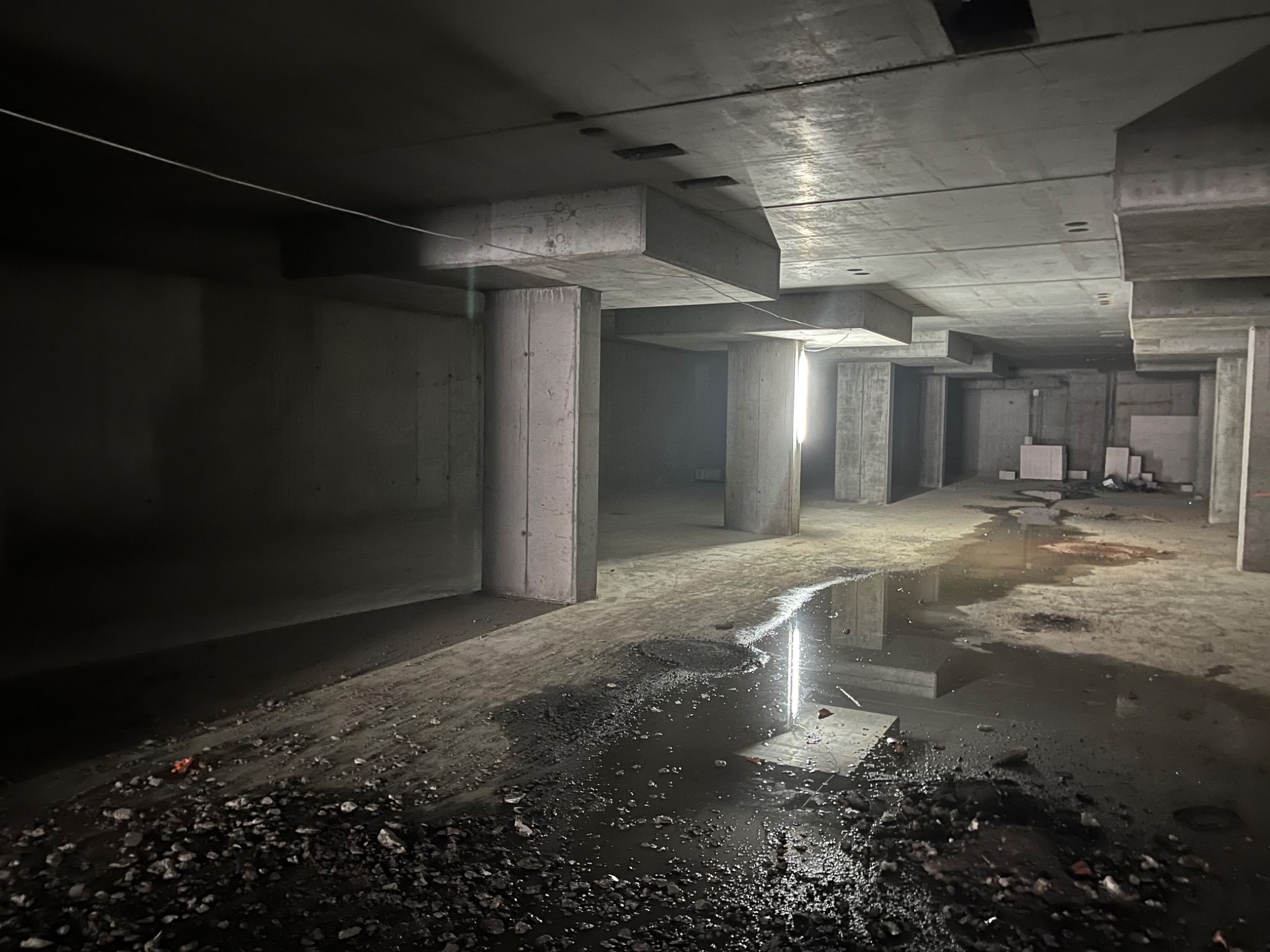 Surowy, betonowy szkielet budynku w trakcie budowy z widocznymi kolumnami, stropami i kałużami na podłodze.