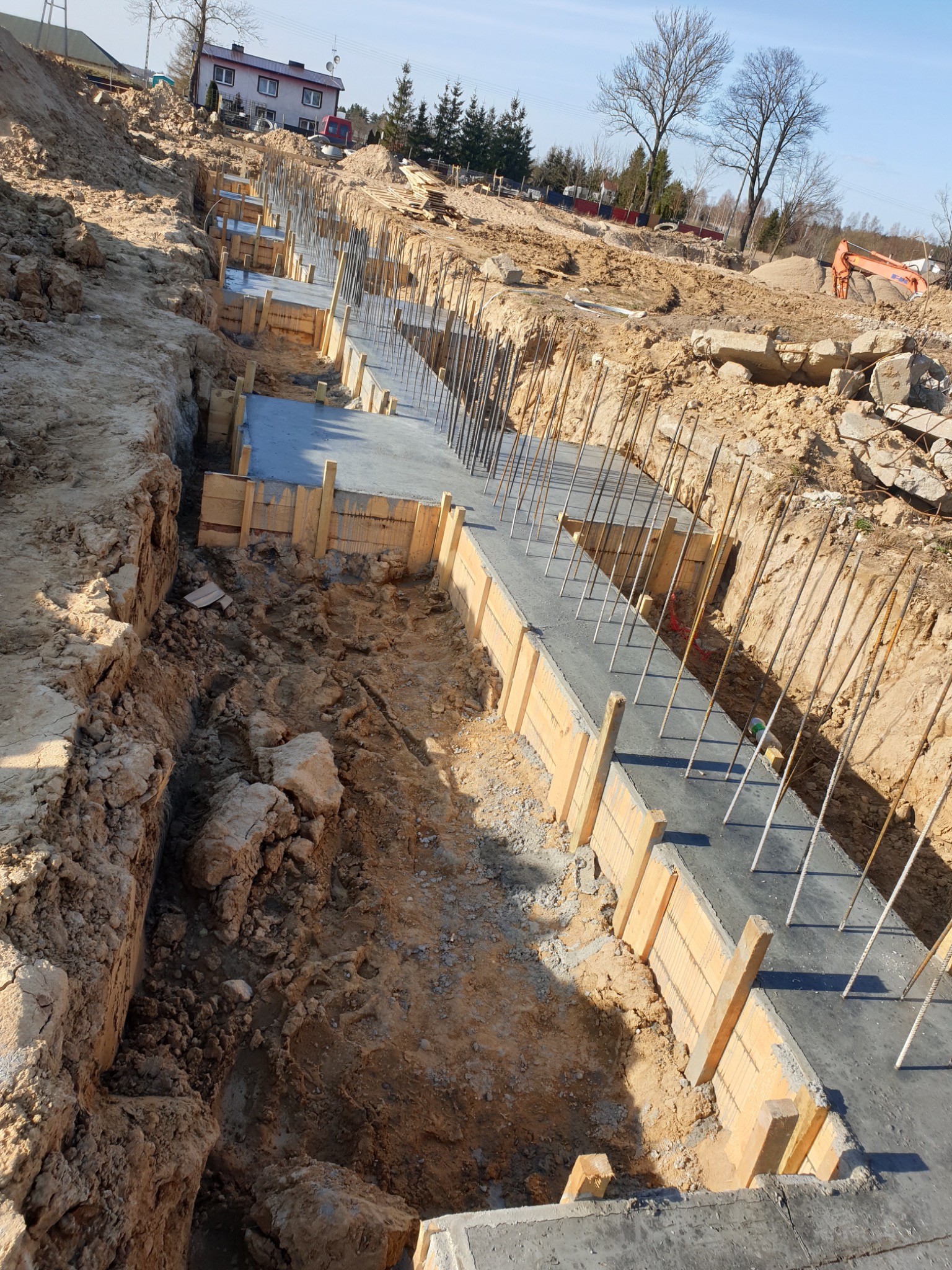 Wykop pod fundament z drewnianym szalunkiem i wystającymi prętami zbrojeniowymi, widok z góry na plac budowy w słoneczny dzień.