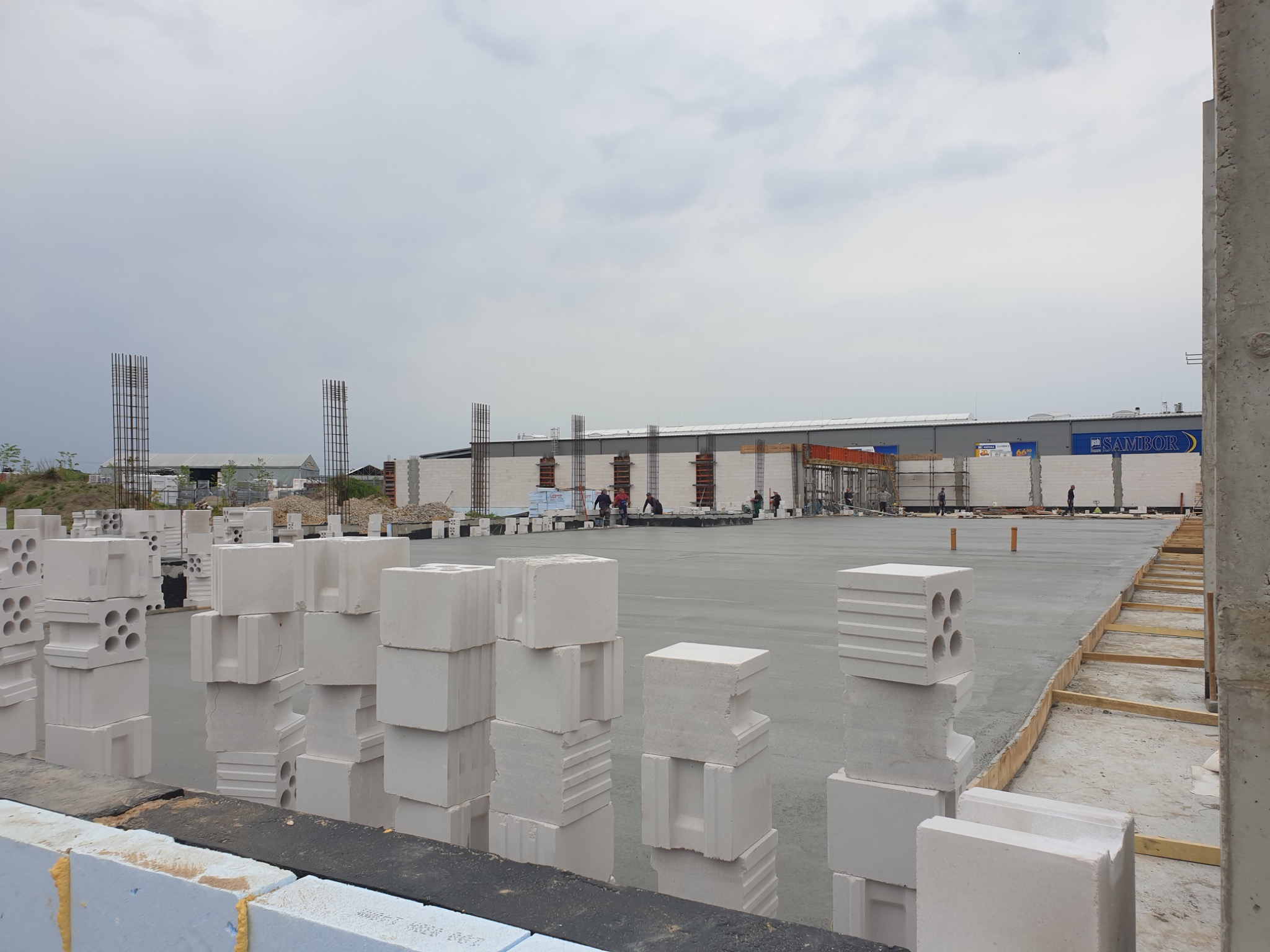 Budowa fundamentów pod budynek w Warszawie, widoczne bloczki betonowe i zbrojenia pionowe, zalana płyta fundamentowa, pracownicy w oddali.