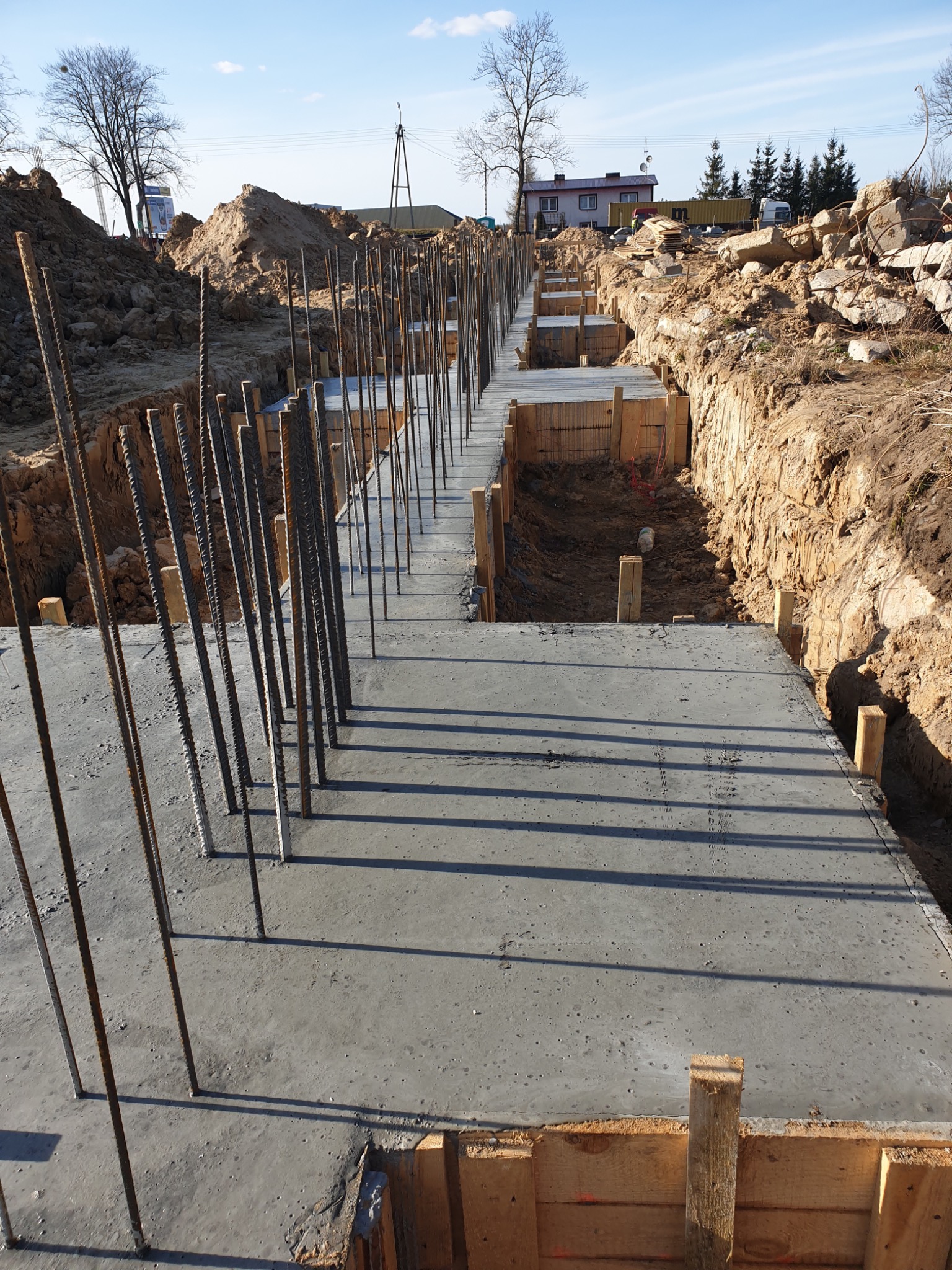 Wylany beton fundamentowy z wystającymi prętami zbrojeniowymi i drewnianym szalunkiem, widok na wykop z ziemią i gruzem oraz budynki w tle.