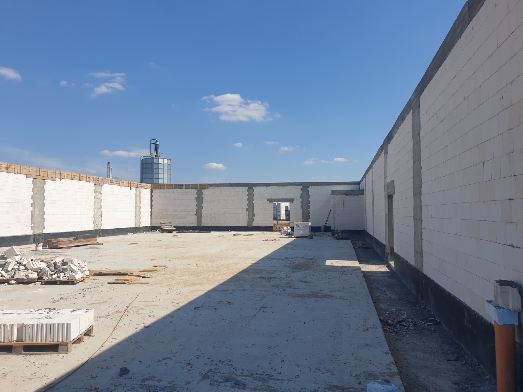 Wnętrze surowego budynku z białych bloczków, widoczne ściany w trakcie wznoszenia, betonowa podłoga z rozrzuconymi materiałami budowlanymi i zbiornik w tle pod błękitnym niebem.