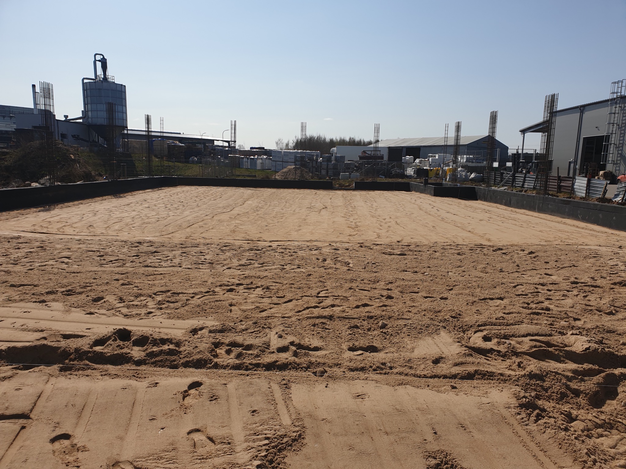 Przygotowany teren pod fundament z widocznymi zbrojeniami i śladami opon pojazdów budowlanych na piasku.