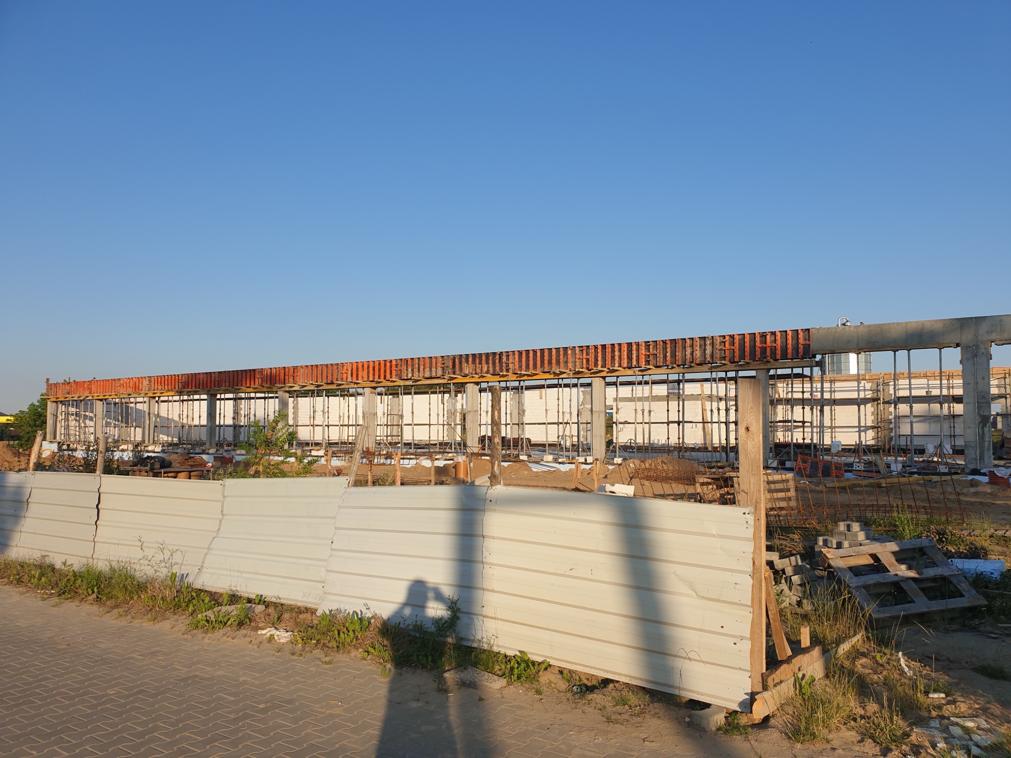 Konstrukcja żelbetowa w trakcie budowy, widoczna spod tymczasowego ogrodzenia z blachy trapezowej, w tle błękitne niebo.
