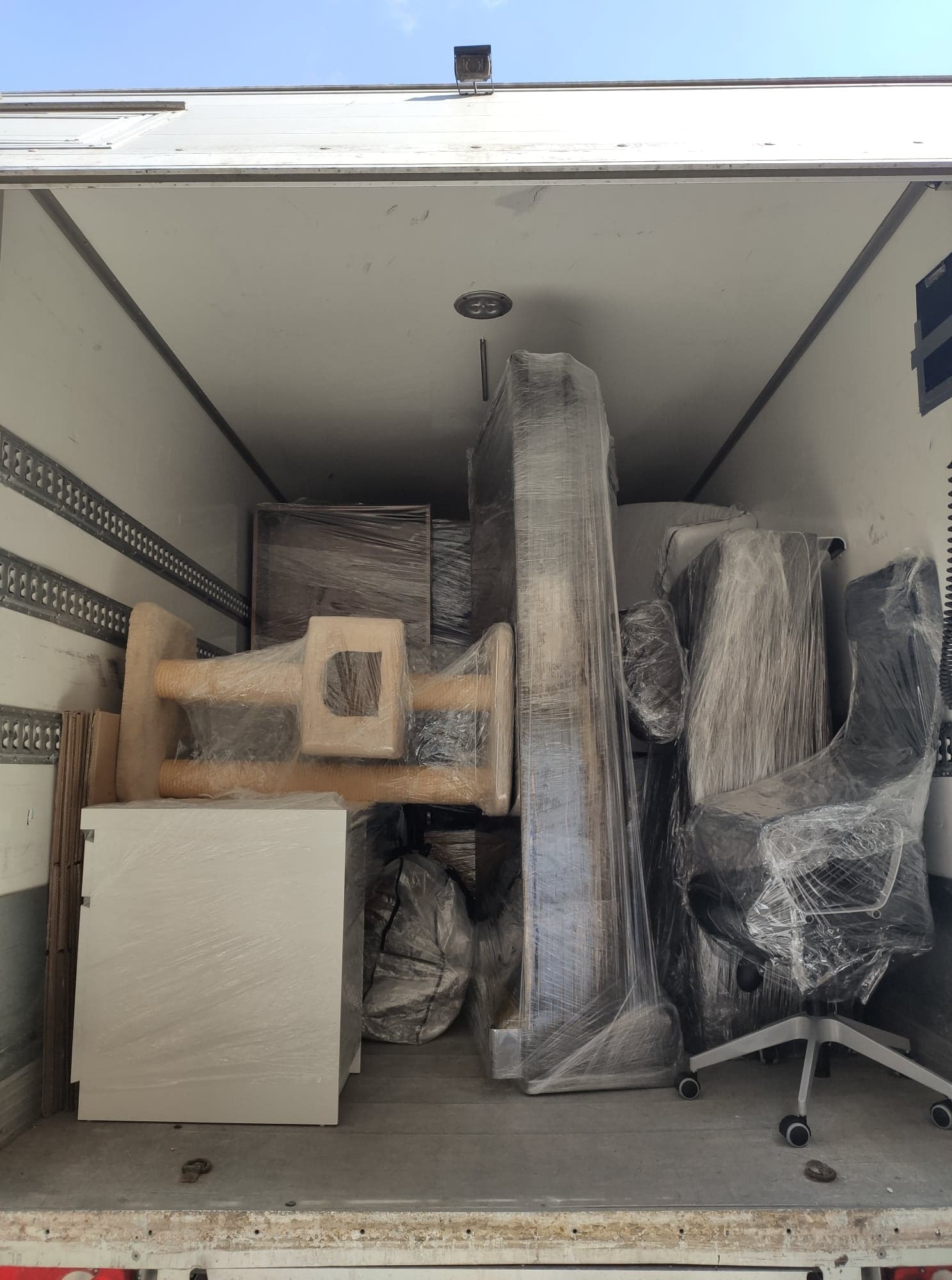 Wnętrze przestrzeni ładunkowej wypełnione meblami owiniętymi folią stretch, widoczne biurko, krzesło obrotowe i inne elementy wyposażenia, zabezpieczone do transportu.