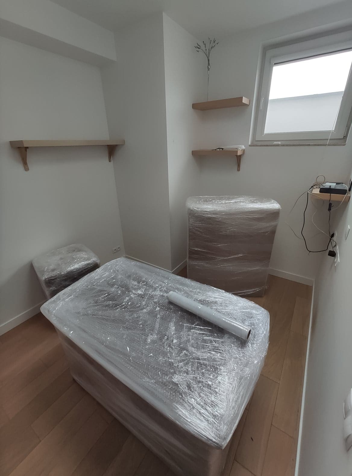 Meble owinięte folią stretch przygotowane do transportu w jasnym pomieszczeniu z drewnianą podłogą i półkami na ścianach.