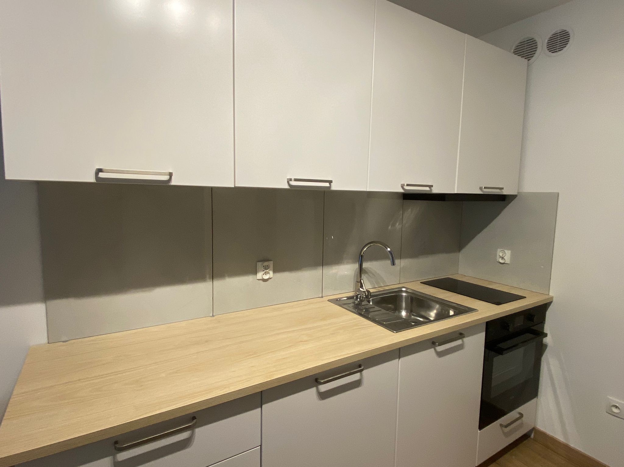 Nowoczesna, minimalistyczna kuchnia z białymi szafkami, drewnianym blatem, stalowym zlewem i płytą indukcyjną. Dwa okrągłe nawiewy wentylacyjne w górnym rogu ściany.