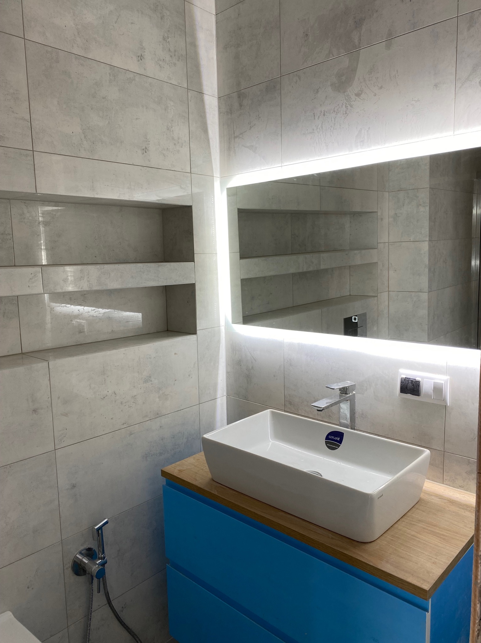 Nowoczesna łazienka z umywalką nablatową na niebieskiej szafce z drewnianym blatem, podświetlane lustro i półki w ścianie wyłożonej szarymi płytkami.